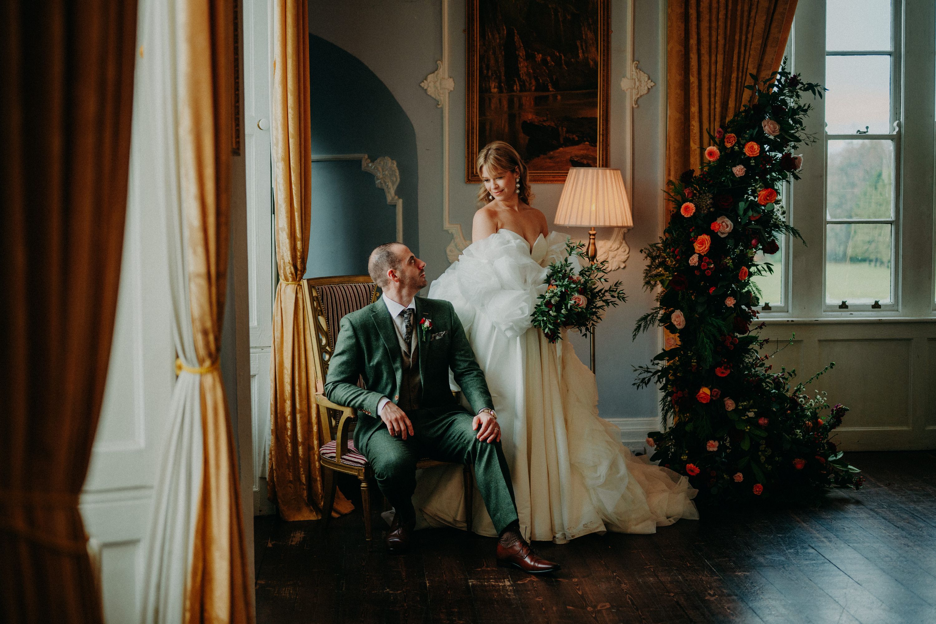 Bride in a strapless tulle gown perches on her groom's lap in a gold-curtained room, a rose arch glowing beside them at Kinnitty Castle, Ireland.