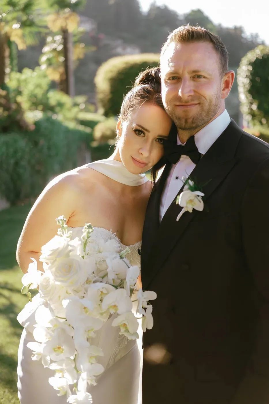 Bride and groom share a close embrace in a sunlit Italian garden with clipped hedges, her cascading orchid bouquet between them.