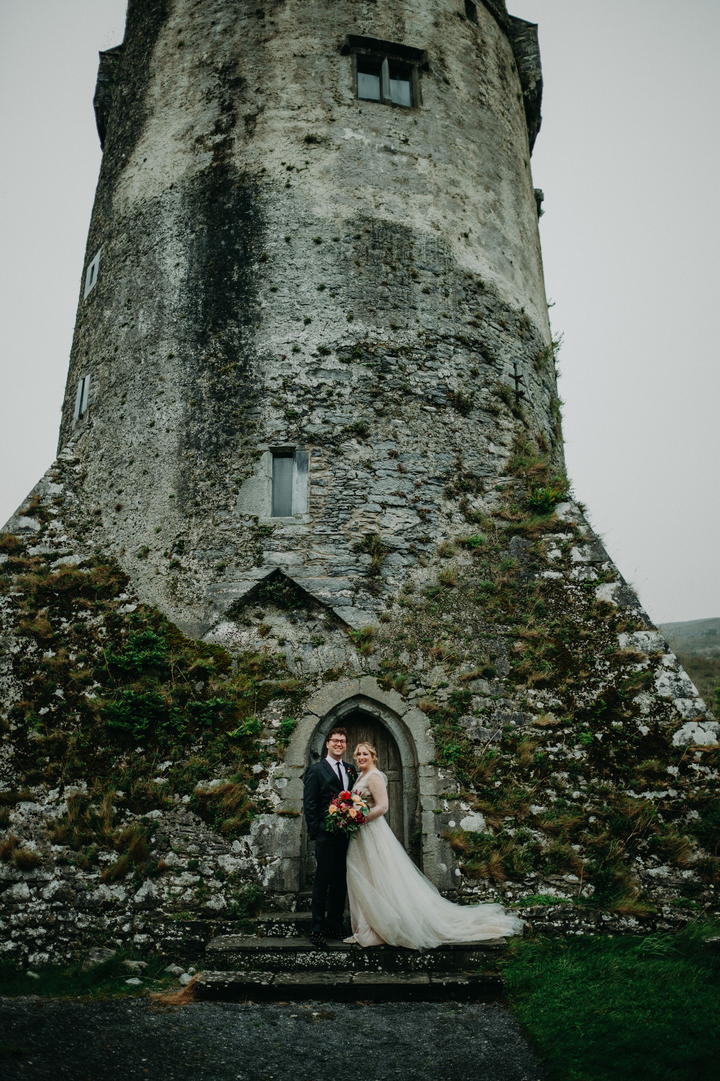 Newlyweds pose beneath a historic stone tower during a small wedding in Ireland with dramatic castle architecture