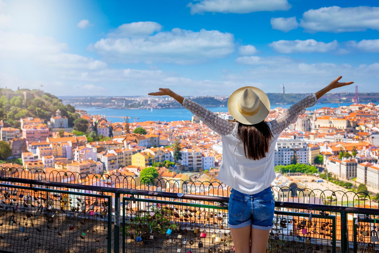 A woman with a hat raising her hands in celebration while looking at the picturesque Portuguese cityscape