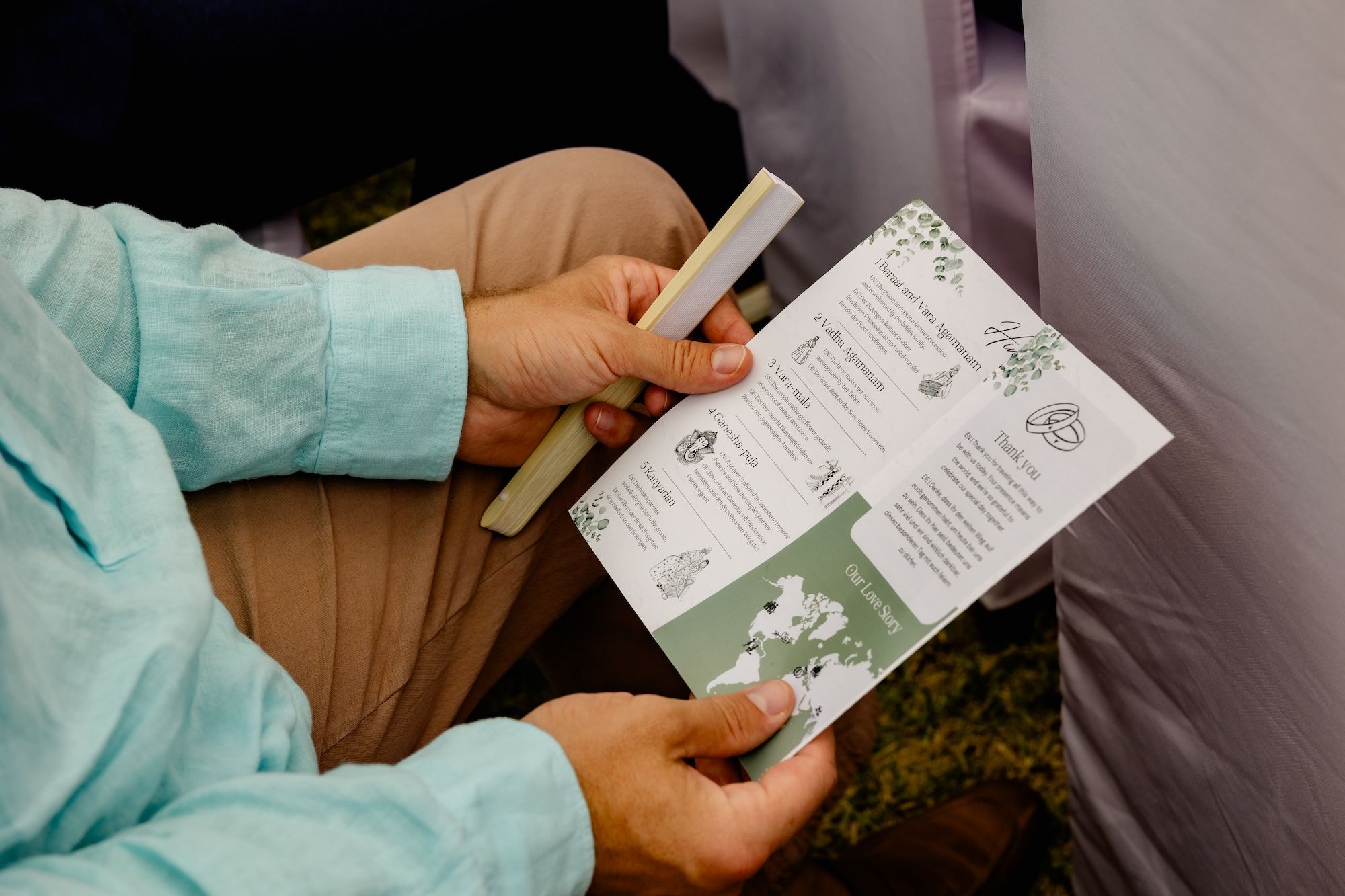 A man in blue long sleeves and brown pants, holding a wedding program and a hand fan