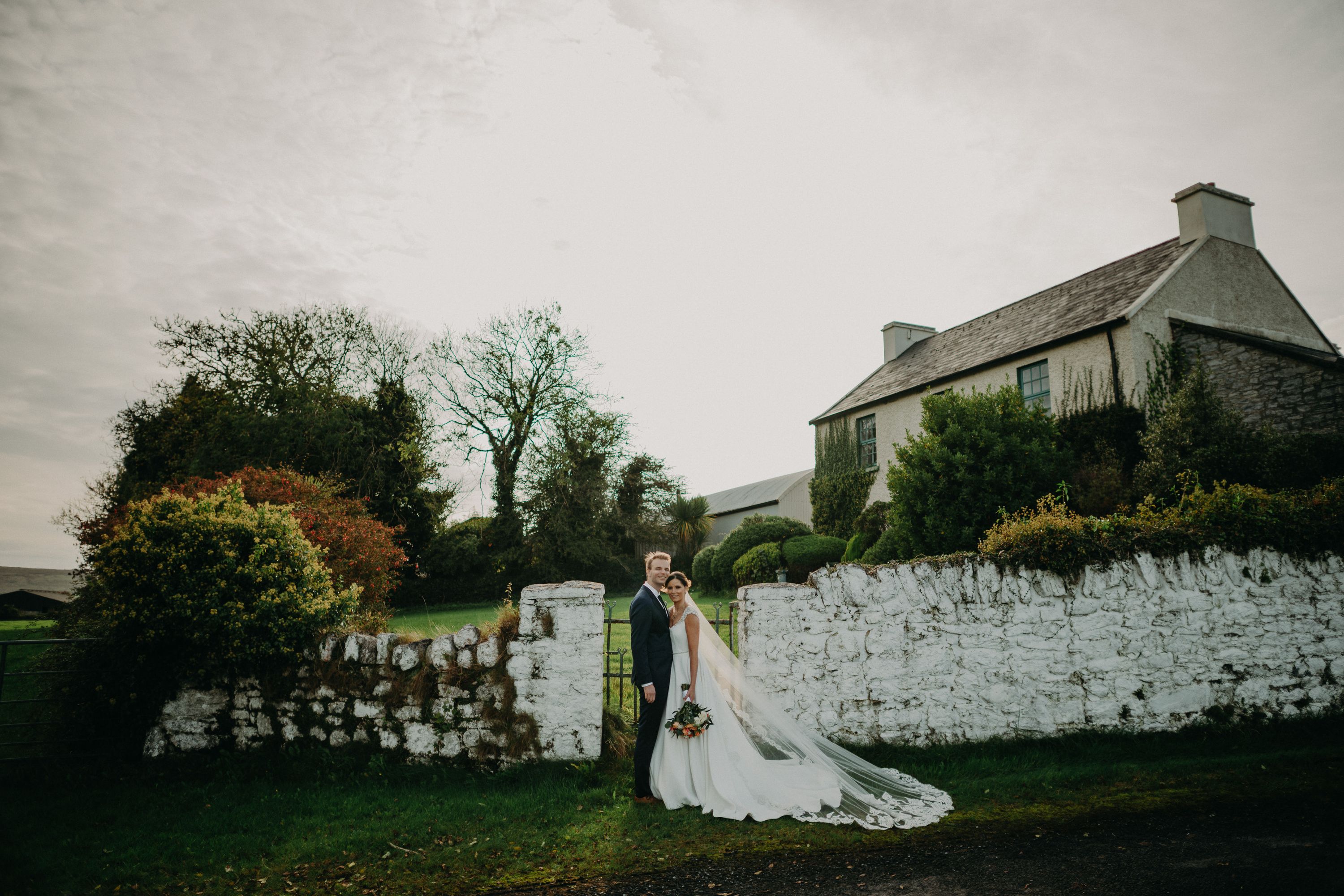 Small wedding in Ireland couple standing outdoors in the garden near stone walls under an overcast sky.