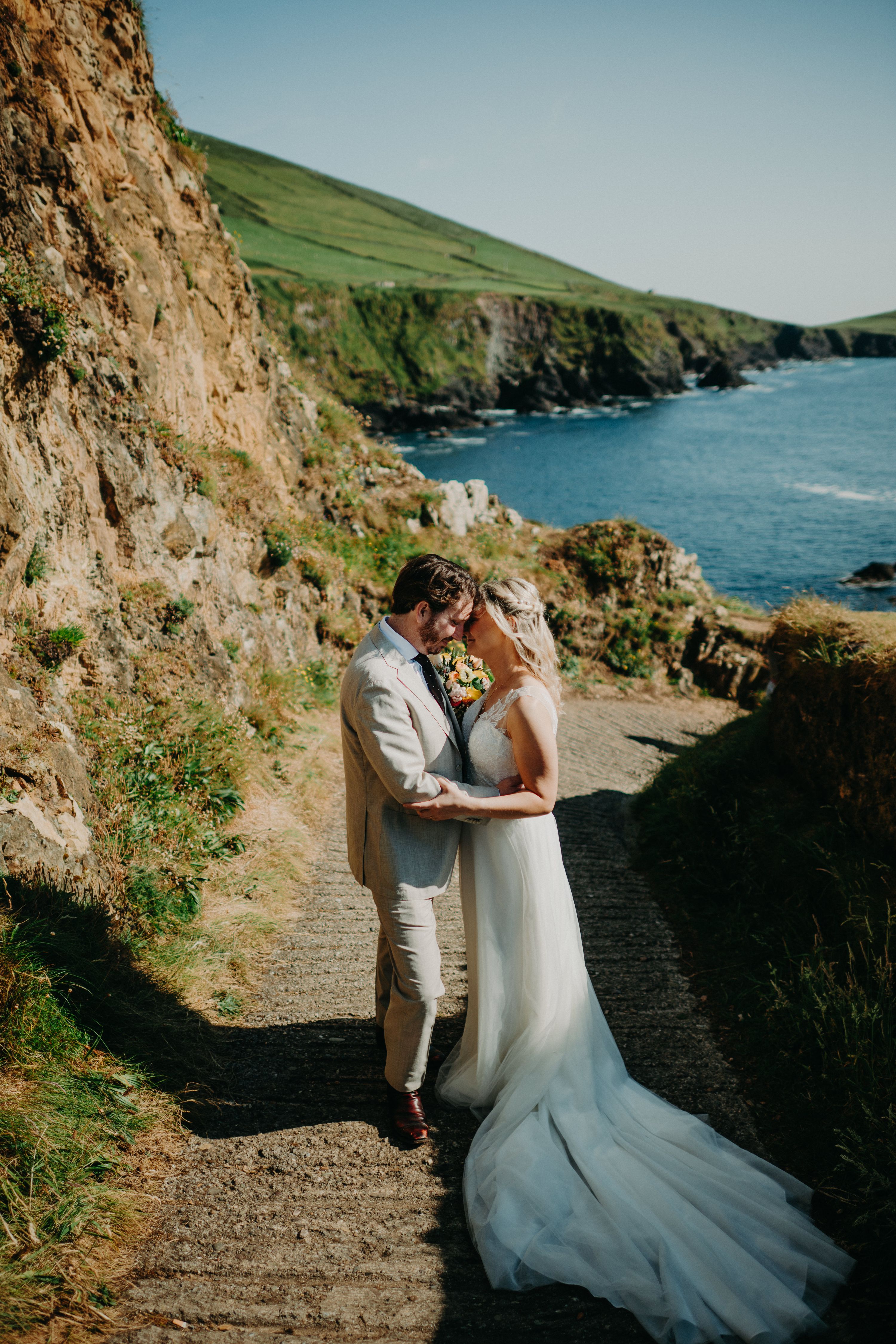 Bride in a tulle A-line gown and groom in a beige suit touch foreheads on the concrete path at Dunquin Pier, green cliffs and blue sea behind.