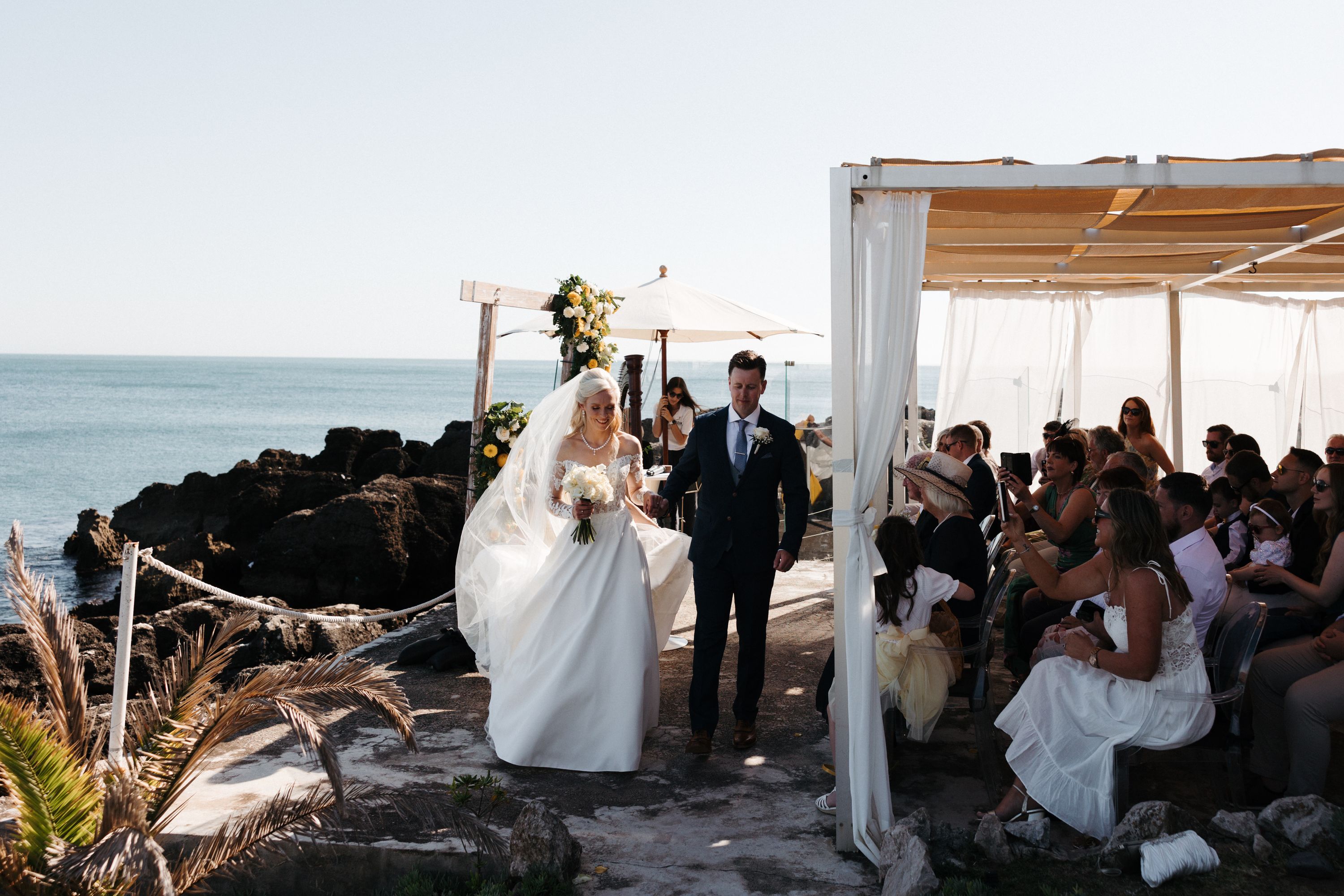 Bride and groom walk during the ceremony of their small wedding in Portugal with the Estoril coast in the background