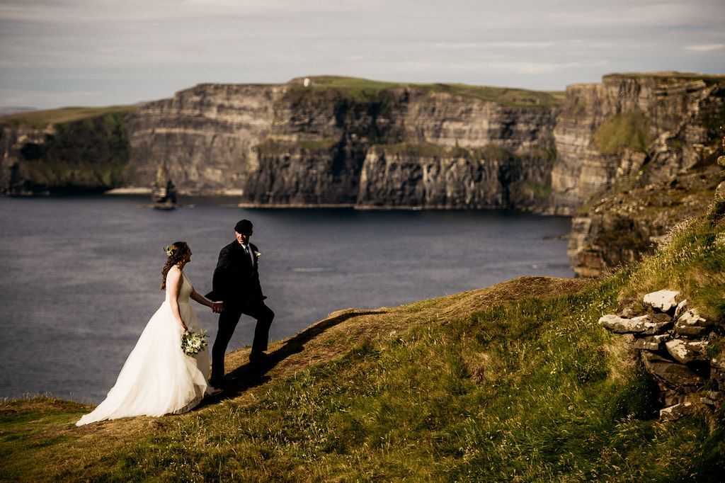 Bride and groom hold hands as they walk up an upward slope on a cliff with Cliffs of Moher and the Atlantic Ocean at the back