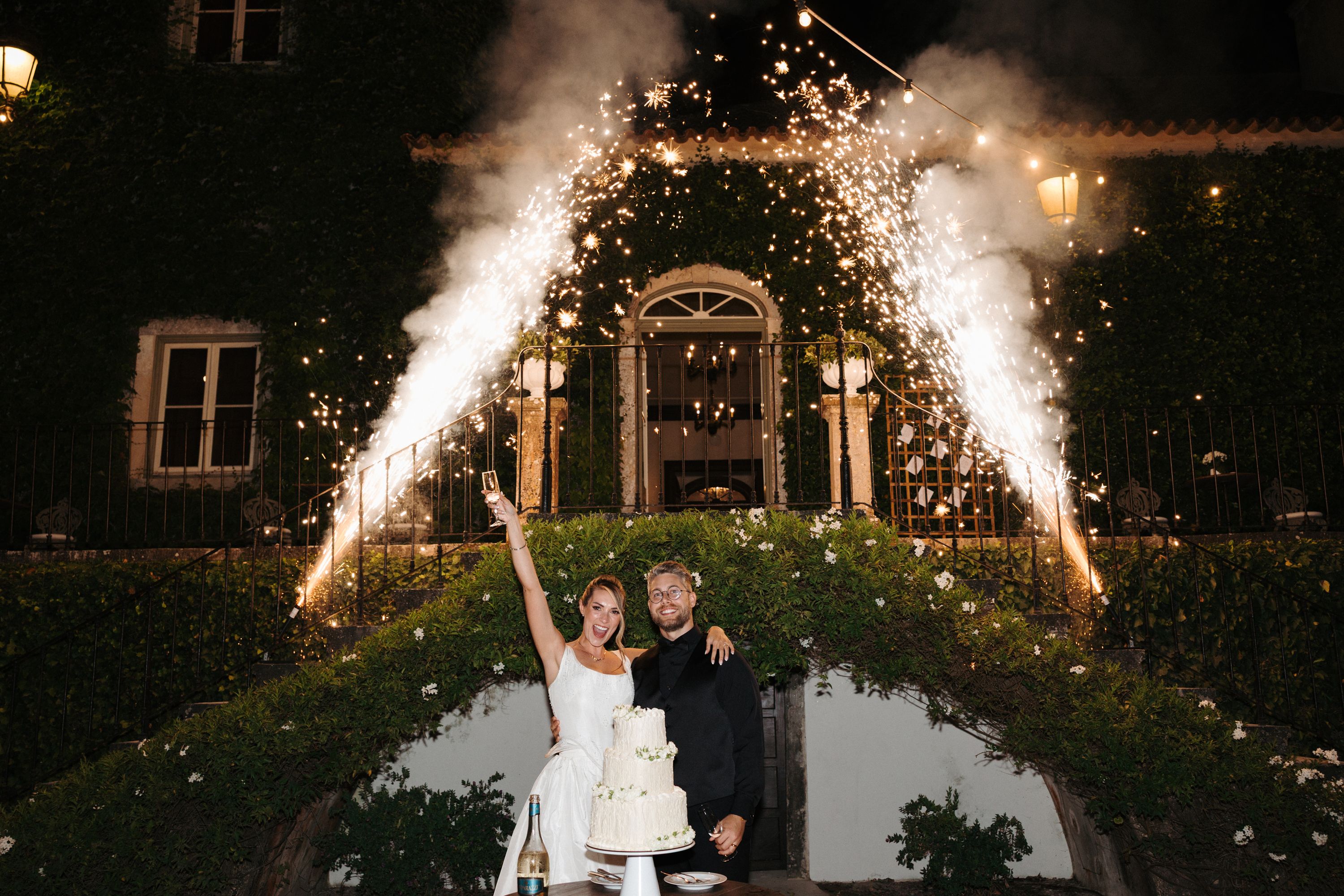 Bride and groom celebrating with a wedding cake and fireworks in the background