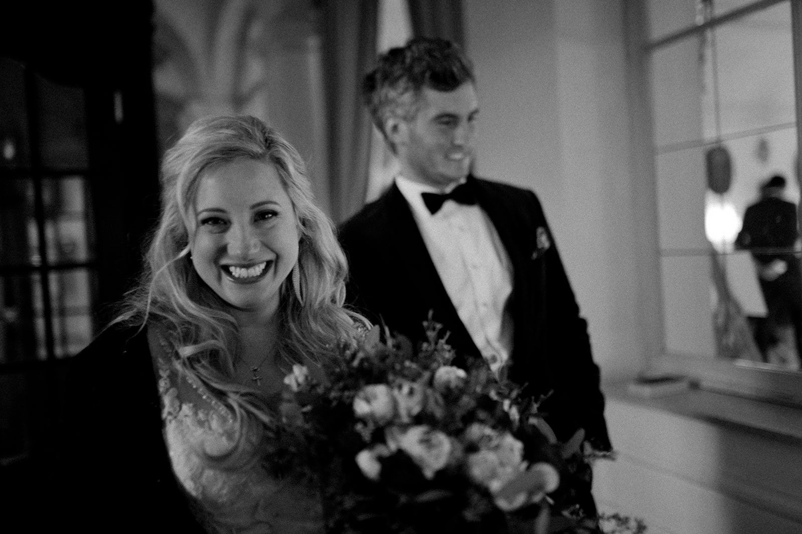 Bride smiling with her bouquet, groom behind her, in black and white