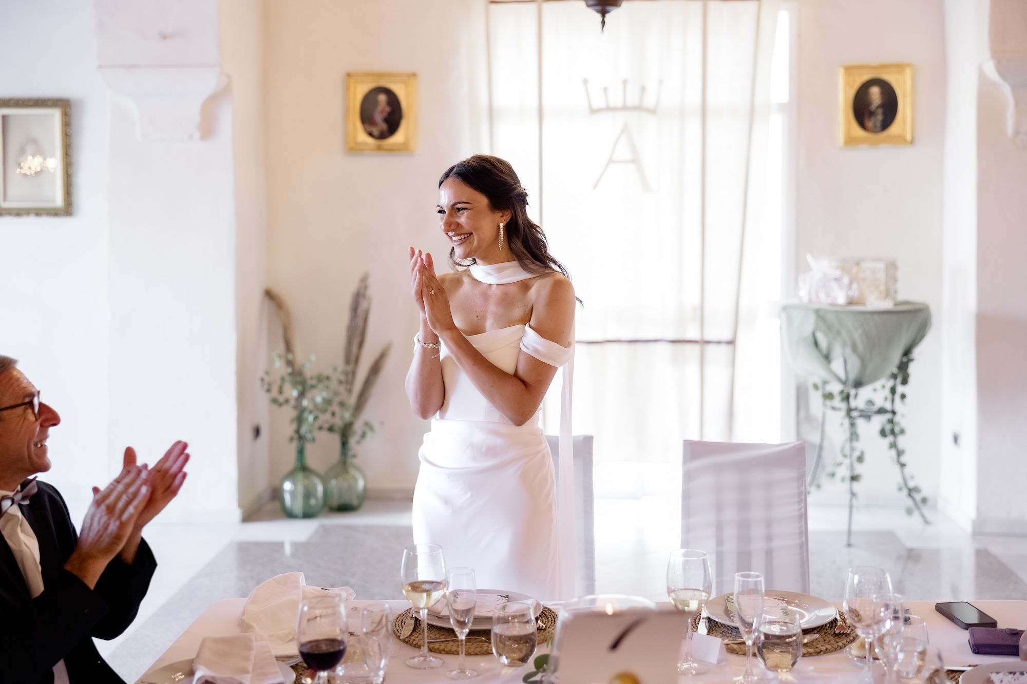 Bride wearing off shoulders dress inside a white-themed event room, laughing and clapping