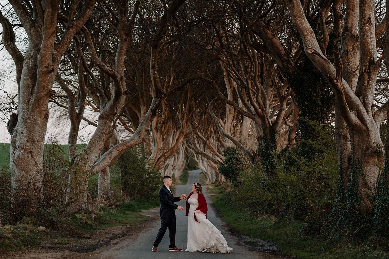 Newlyweds hold each other’s hands in the middle of the road, with a lineup of large old beech trees on both sides