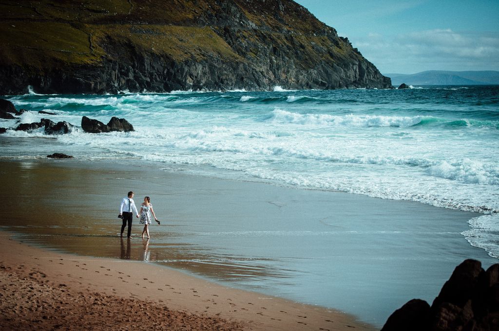 A couple walking along a white-sand beach with ocean waves splashing in the background during their elopement in Ireland