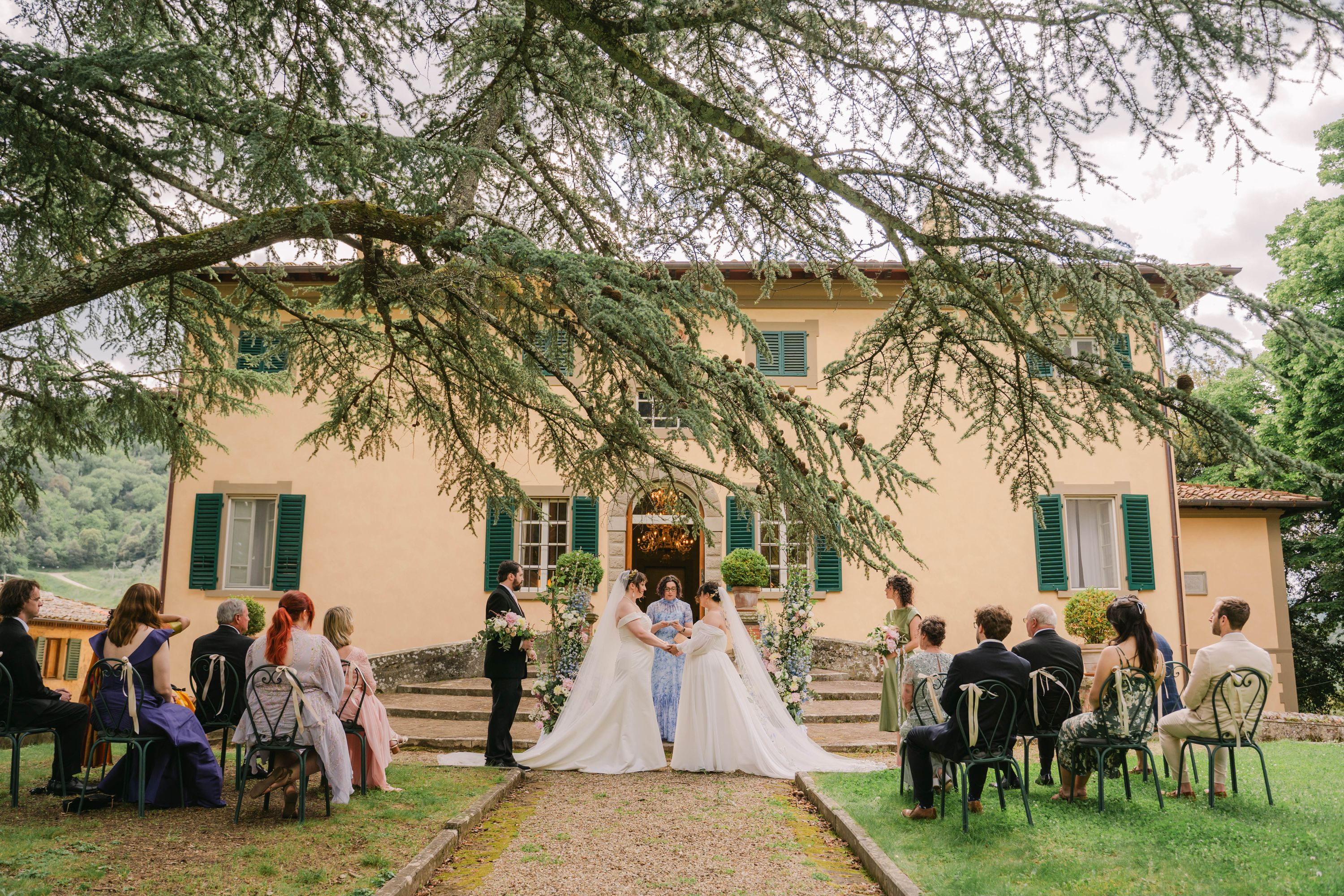 Two brides say their vows on the lawn below the villa steps, around twenty guests seated on either side under a spreading cedar of Lebanon.