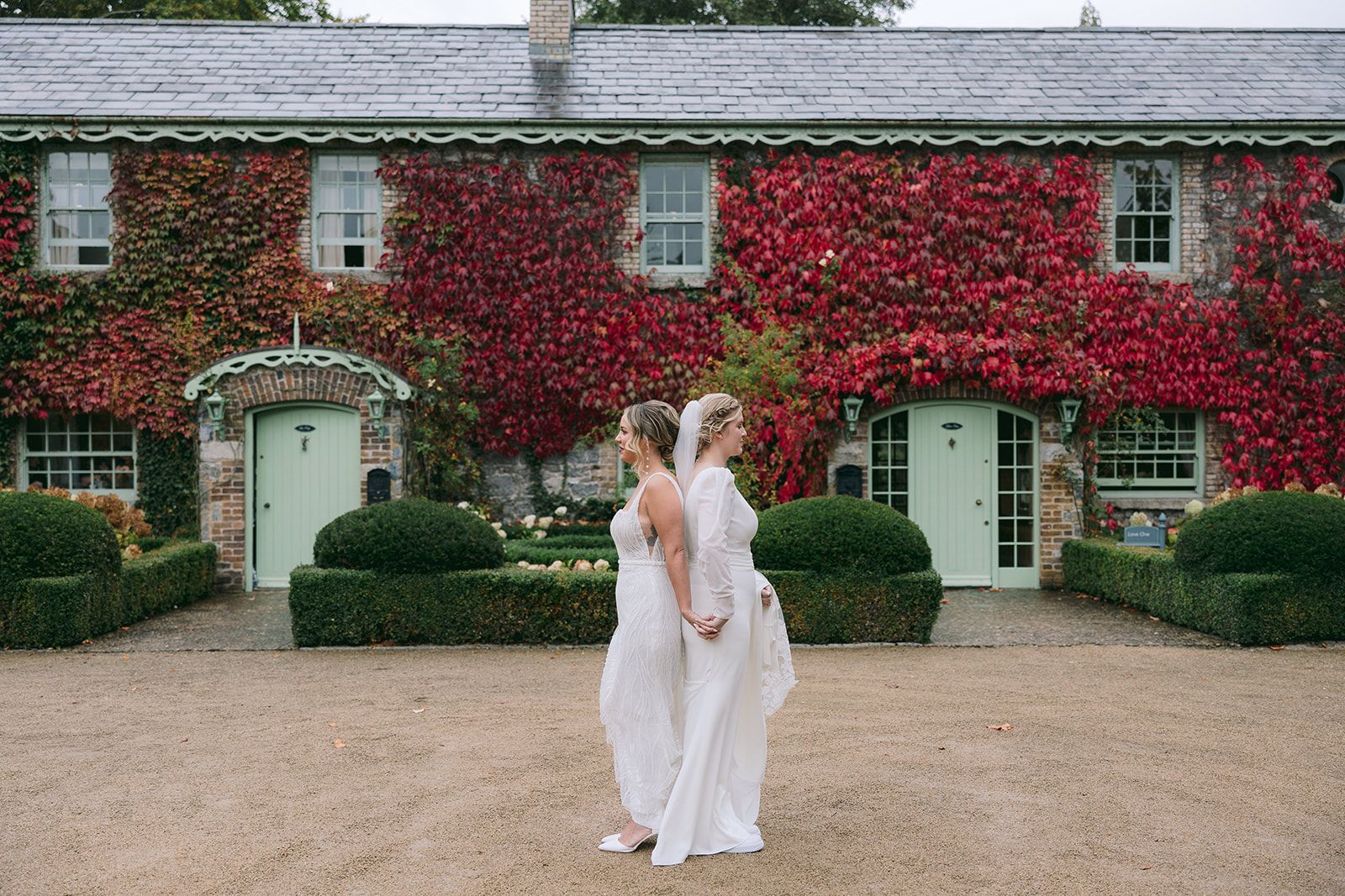 Two brides in wedding dresses back to back holding hands in front of a rustic building during their small wedding in Ireland