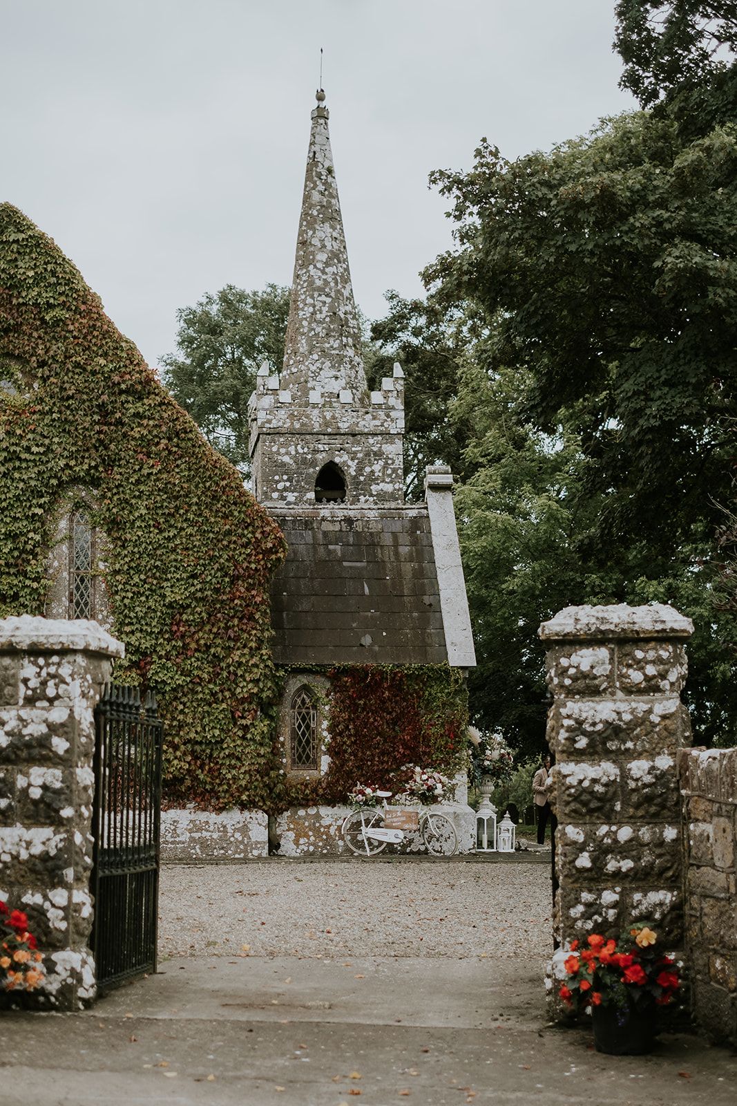 Through the gate piers of St. John's Church, Knockainey: the crenellated bell tower and spire, the creeper-covered gable and a white bicycle on the gravel.