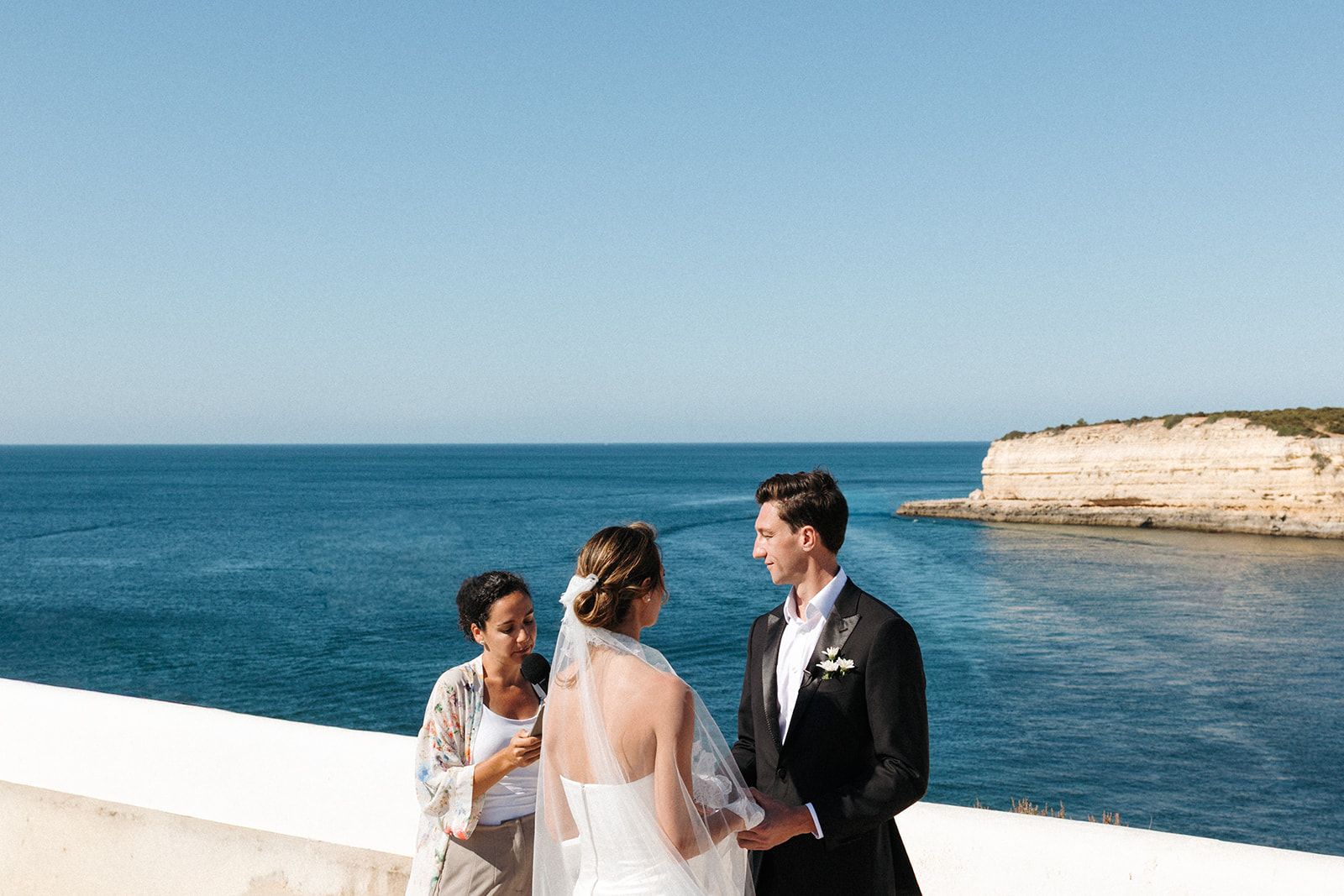 Bride and groom holds hands on a clifftop spot under the clear summer skies of Algarve during their Portuguese wedding abroad