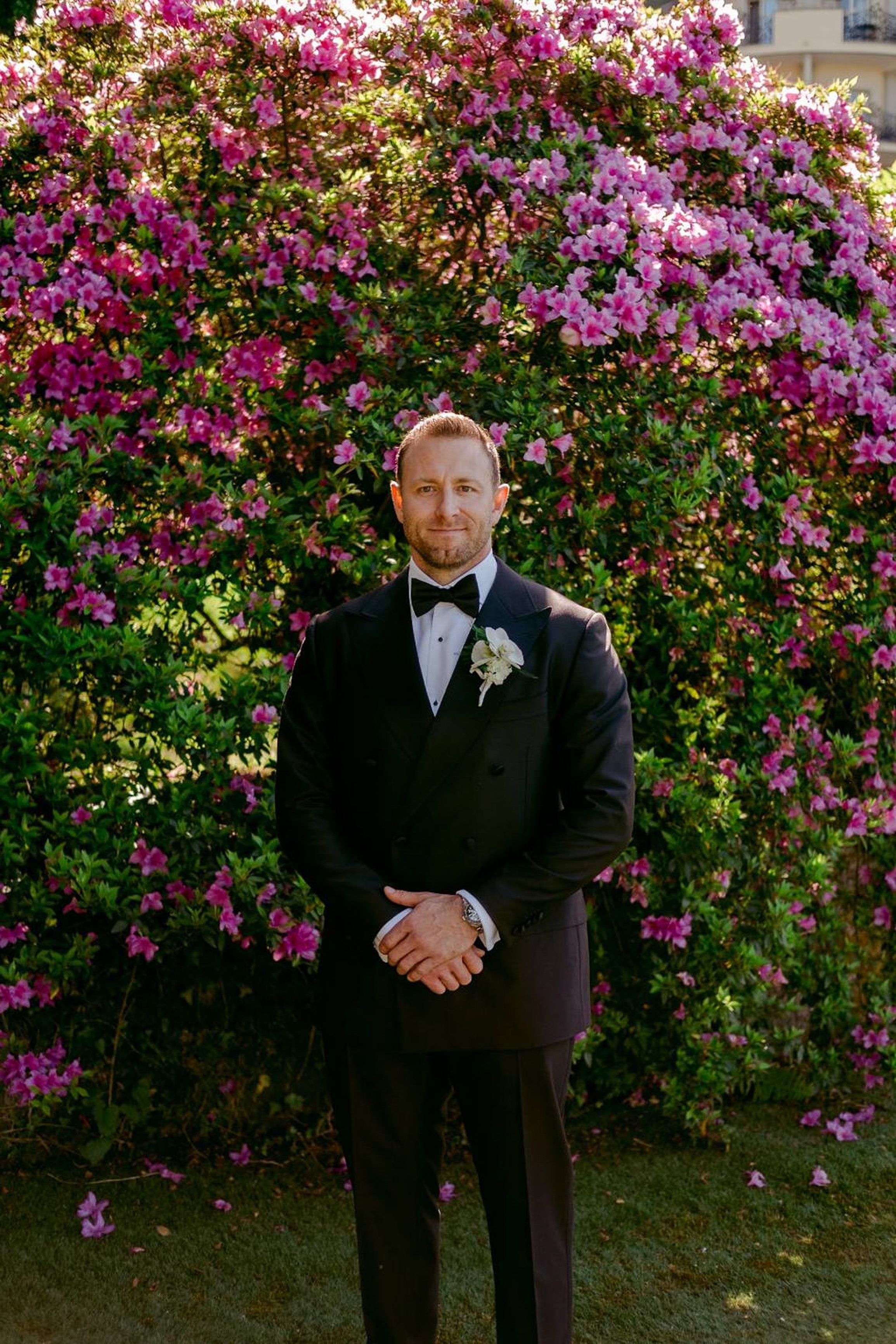 Groom portrait in a black tuxedo standing before blooming pink flowers during an intimate wedding in Italy