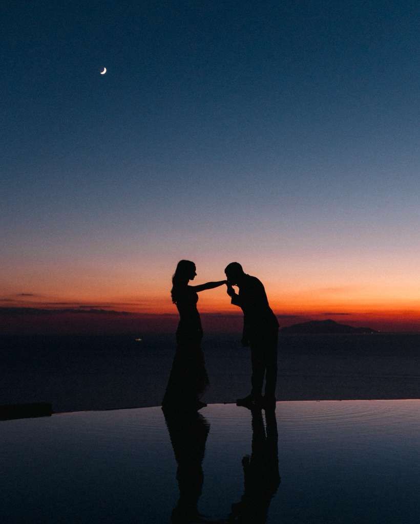 A silhouette of a groom kissing his bride's hands with sunset hues in the background during their micro wedding in Italy