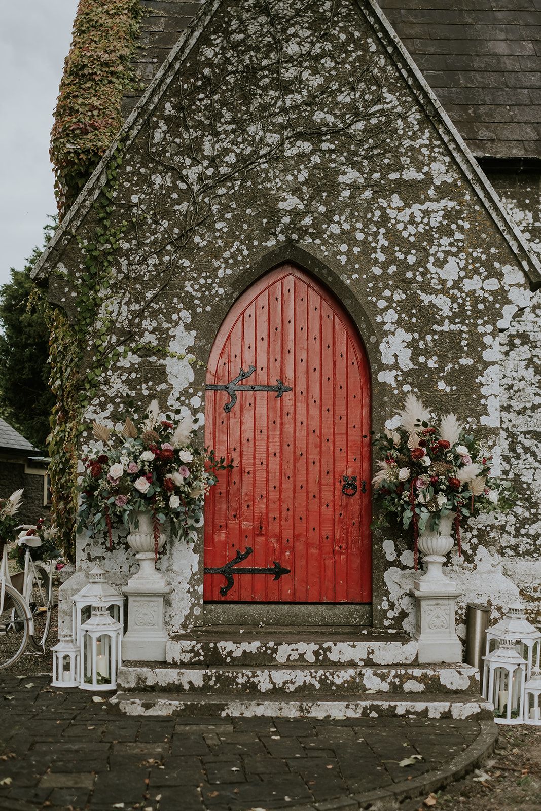Urns of burgundy and blush flowers with pampas grass flank the red studded door of St. John's Church, Knockainey, lanterns on the steps below.
