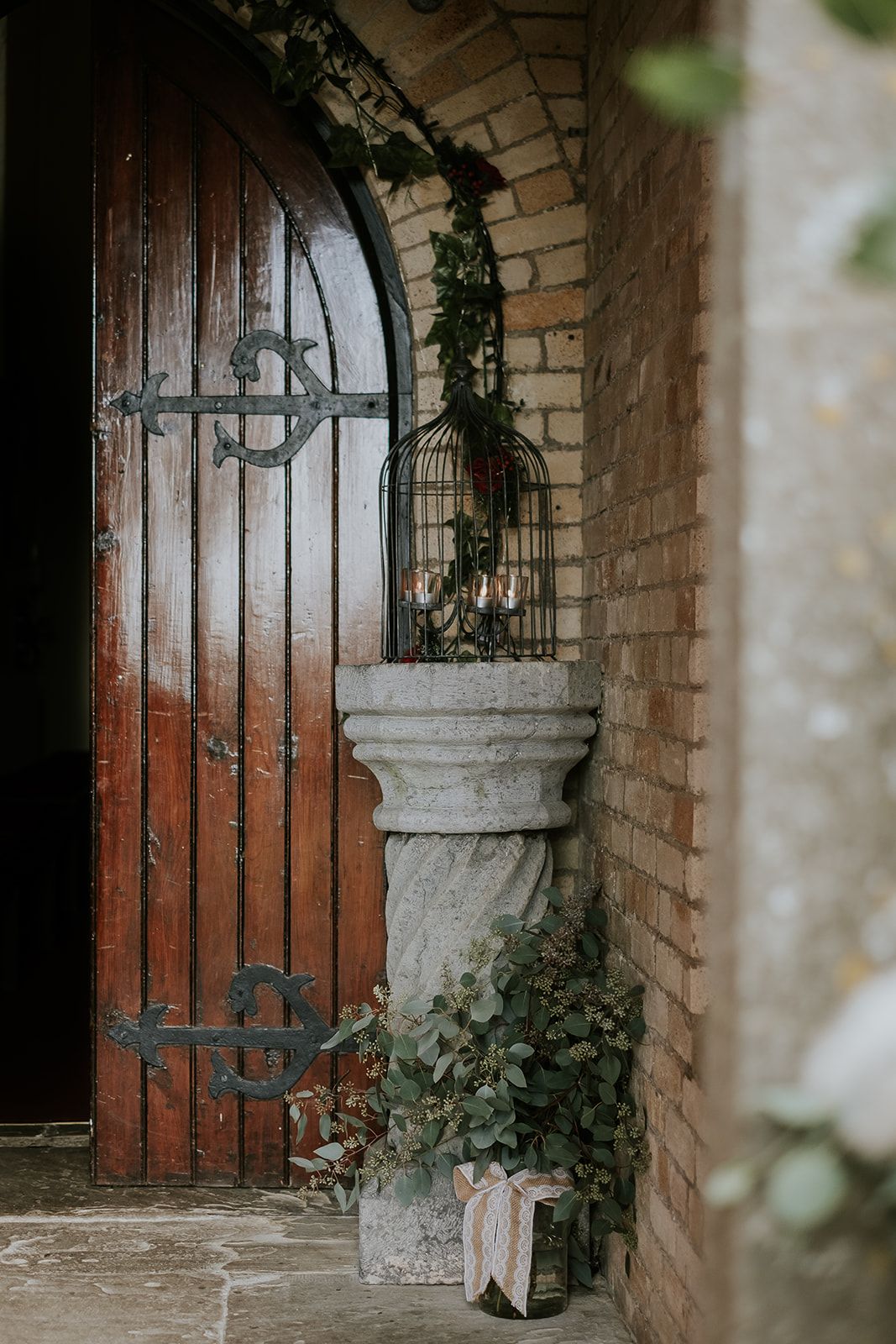 Inside the brick-lined porch of St. John's Church, Knockainey, a birdcage of candles and eucalyptus on a stone pedestal beside the open timber door.