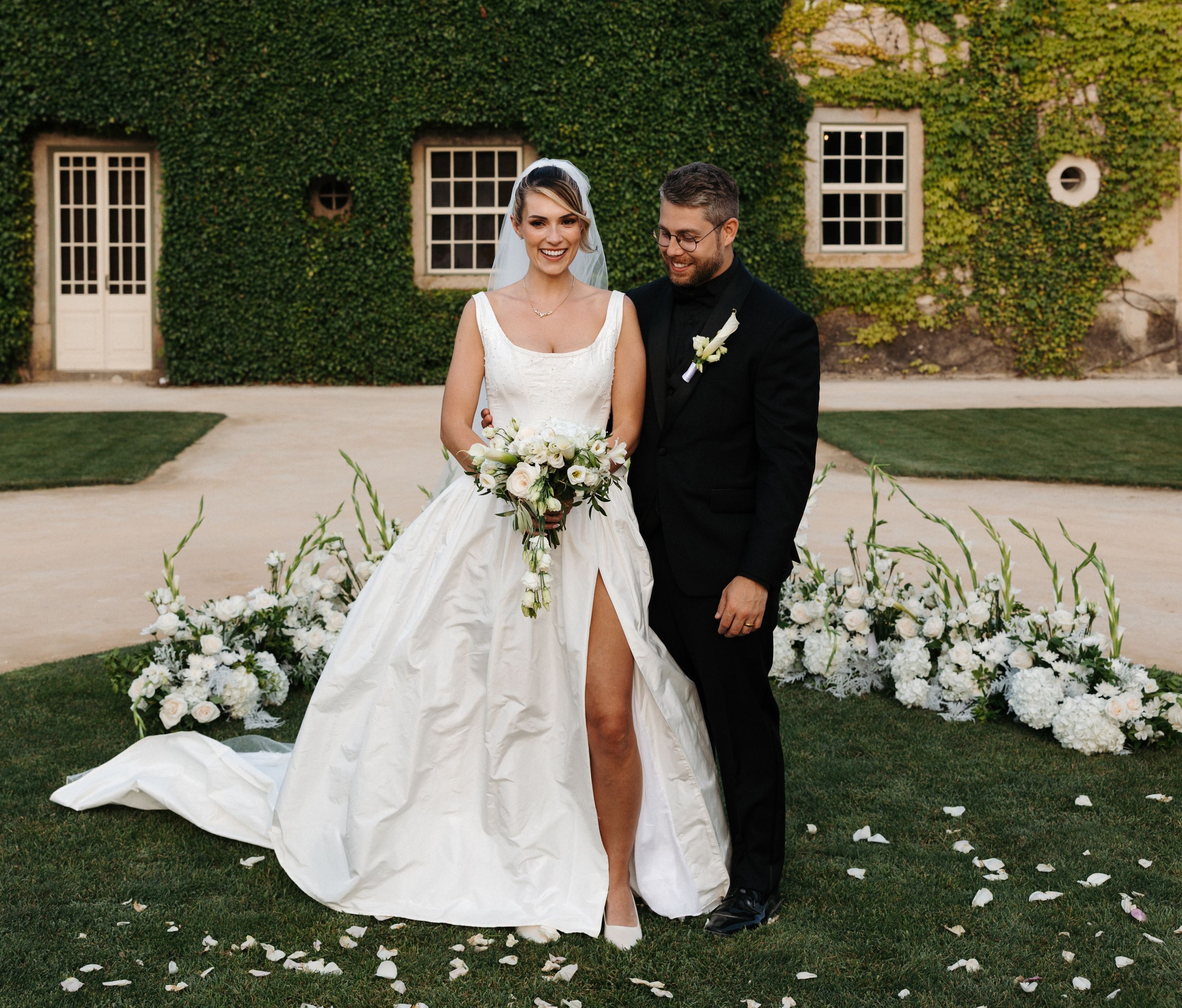 Groom looking at his bride who holds a bouquet in front of an ivy-covered building during their destination wedding in Portugal