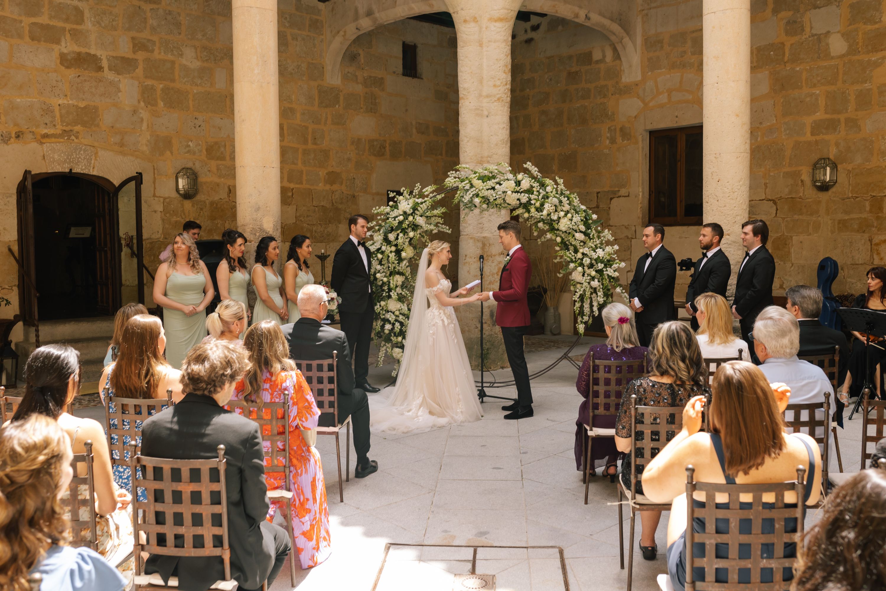 A ceremony in a courtyard with a floral arch, the bride and groom, and entourage in front during a destination wedding in Spain