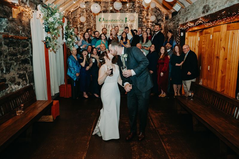 Bride and groom kissing and holding a glass of liquor inside a traditional Irish pub, with their guests in the background