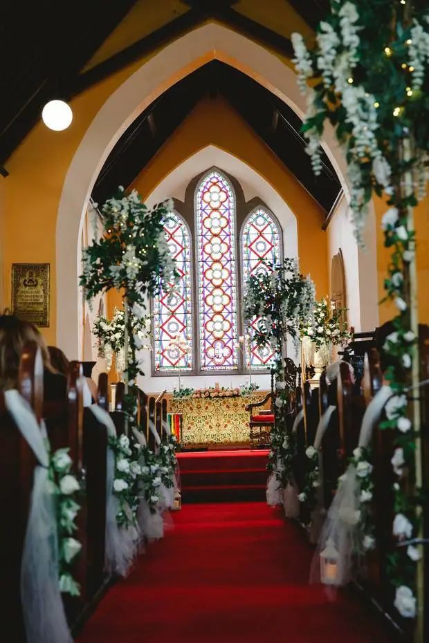 Wedding ceremony inside St. John's Church, Knockainey: a red-carpet aisle lined with white flowers and fairy lights, stained glass beyond the altar.