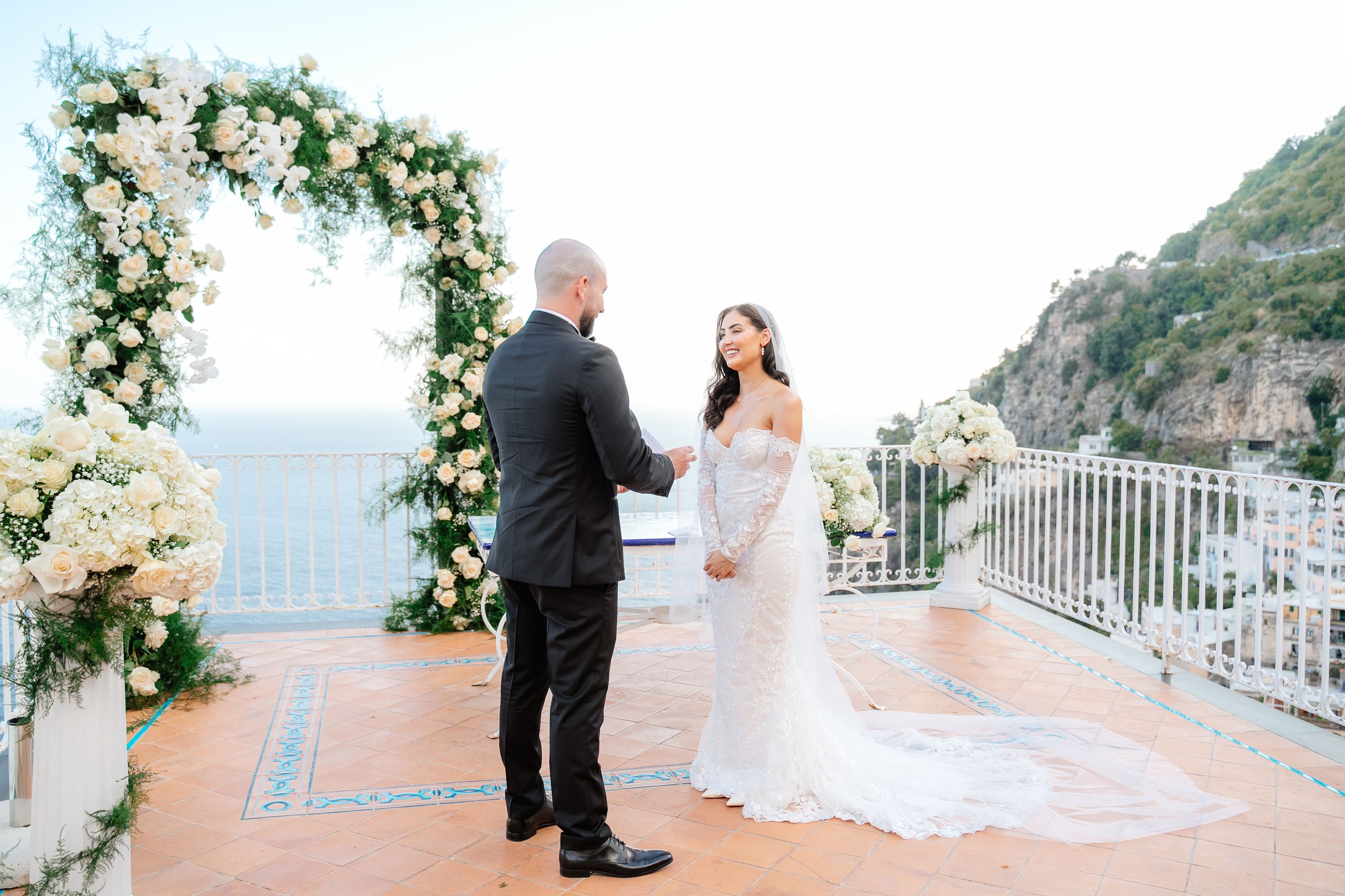 Couple exchanges vows atop a terrace with a floral arch behind them and the view of Amalfi Coast in the background