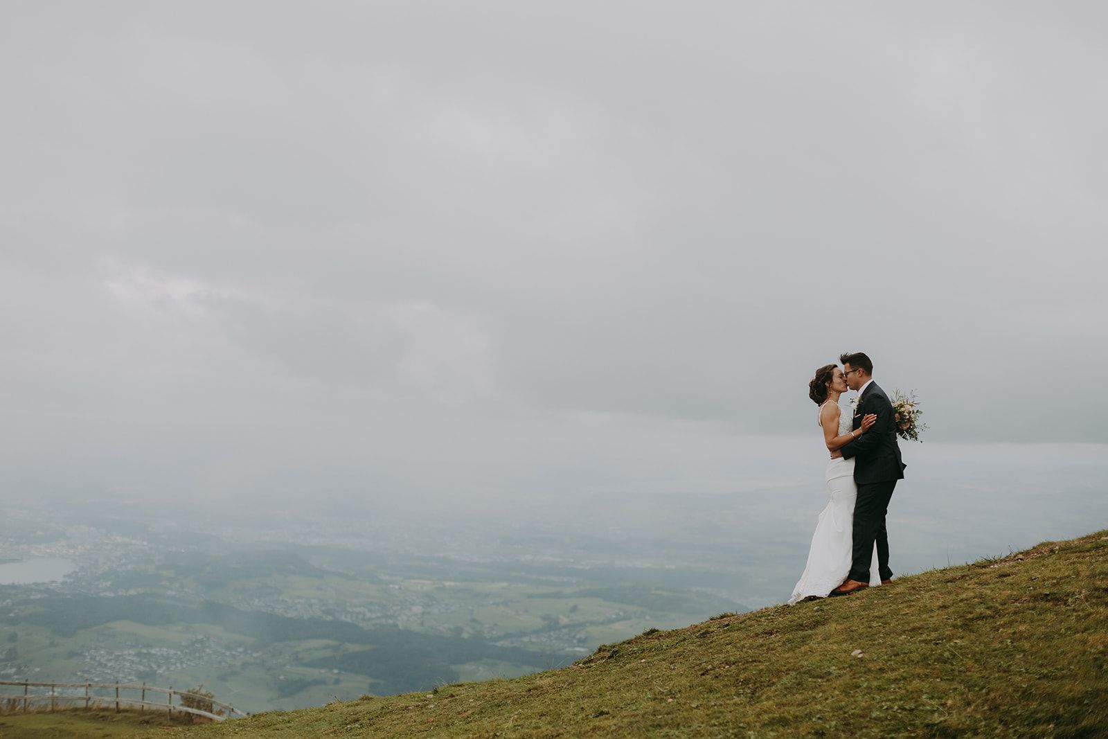 A bride and groom kissing on a hill in front of a foggy backdrop of Swiss scenery