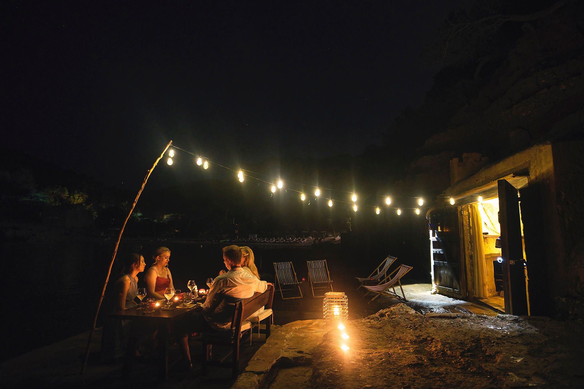 A group of people having dinner outdoors under the night sky with candles on the table and hanging lights above