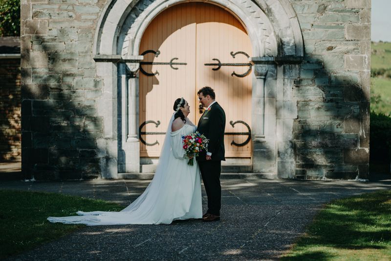 Newlyweds in front of a chapel with a closed wooden door during the photoshoot of their destination wedding in Ireland