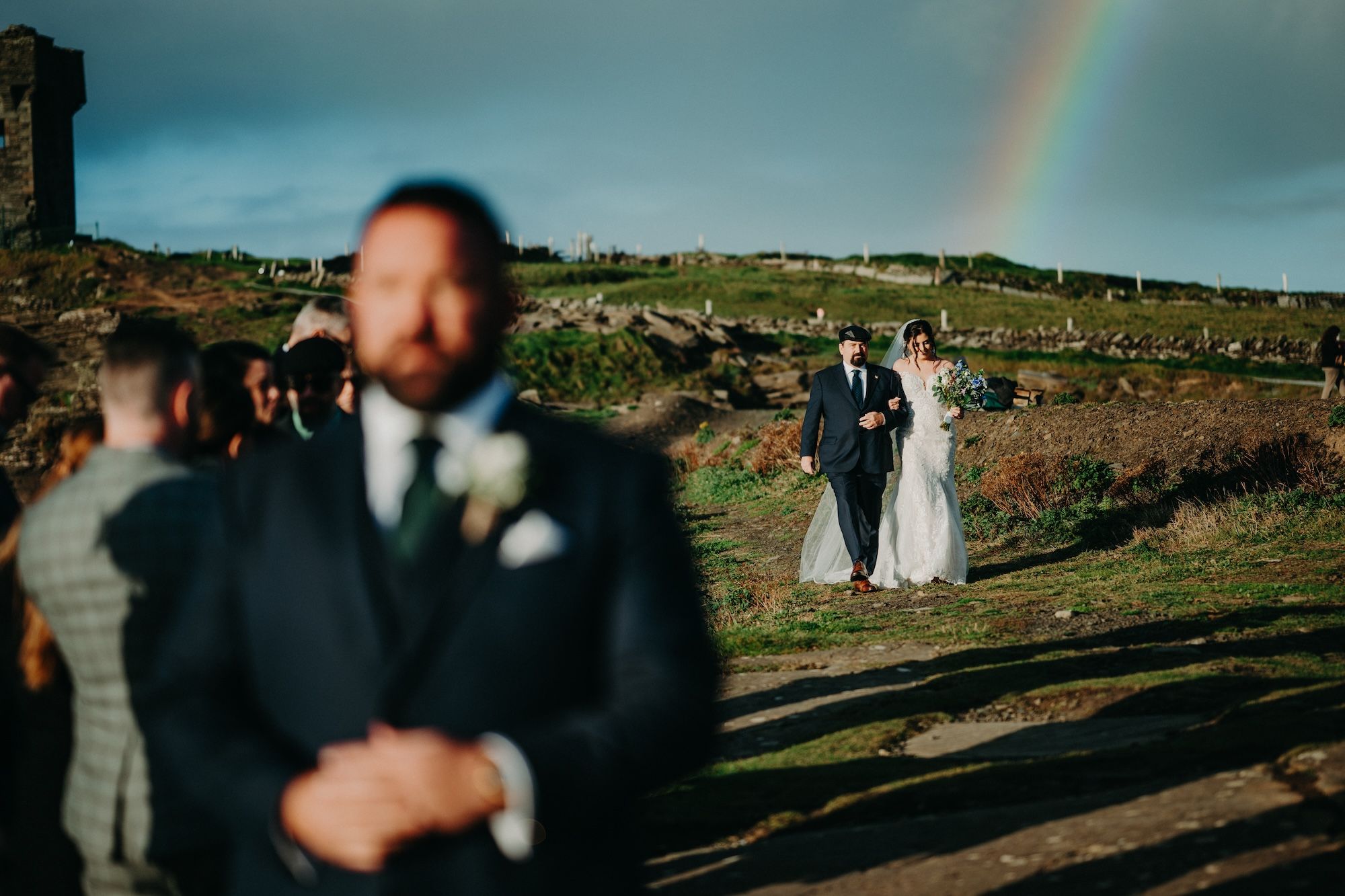 Bride walks down the clifftop aisle with her father with a rainbow in the sky