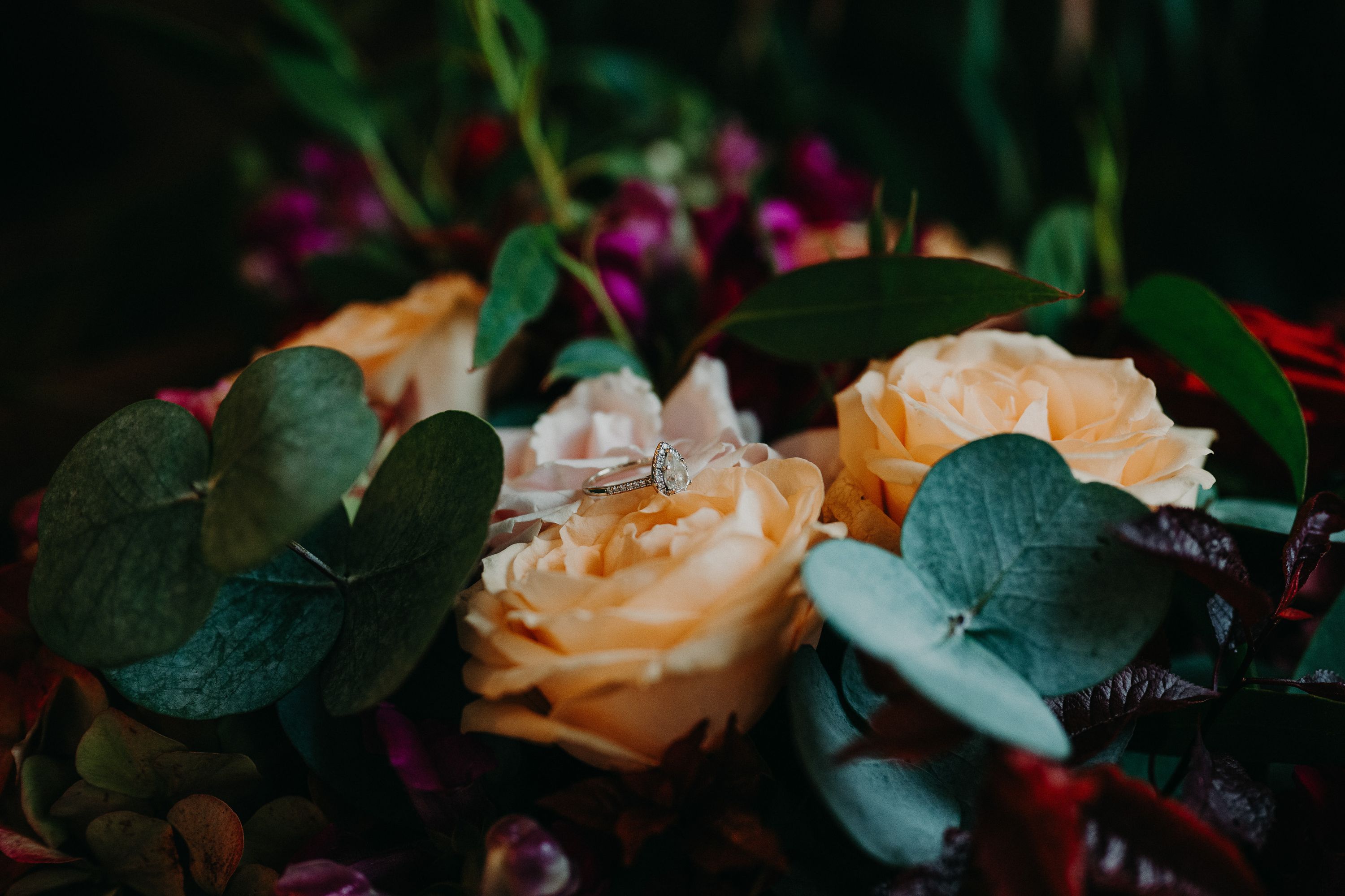 Close-up of a pear-shaped diamond engagement ring resting on peach garden roses and eucalyptus in a bridal bouquet
