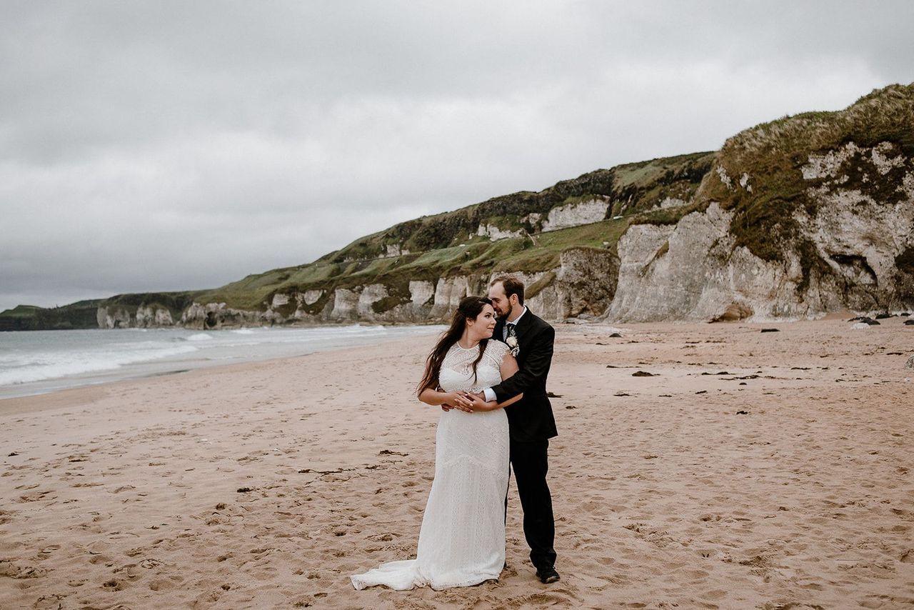 Bride and groom hugging each other on a beach with a sandy shore and white rock formations at the back