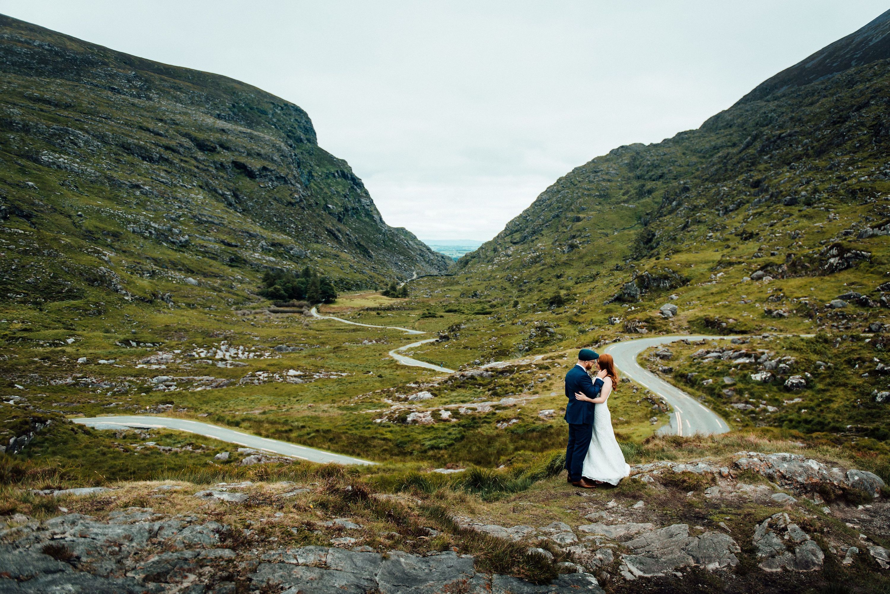 Bride and groom kissing with the winding road, roilling hills, and lush green landscapes of Ireland in the background
