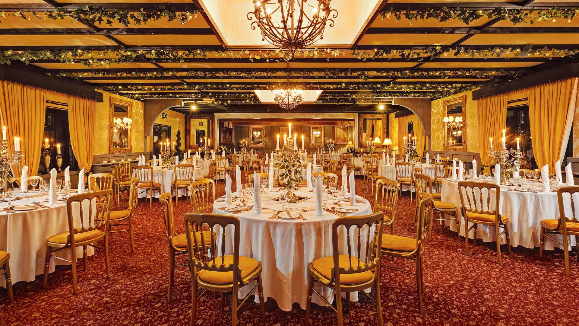 Banqueting hall set for a wedding dinner with round tables, gold chairs, candelabras and ivy-strung beams.
