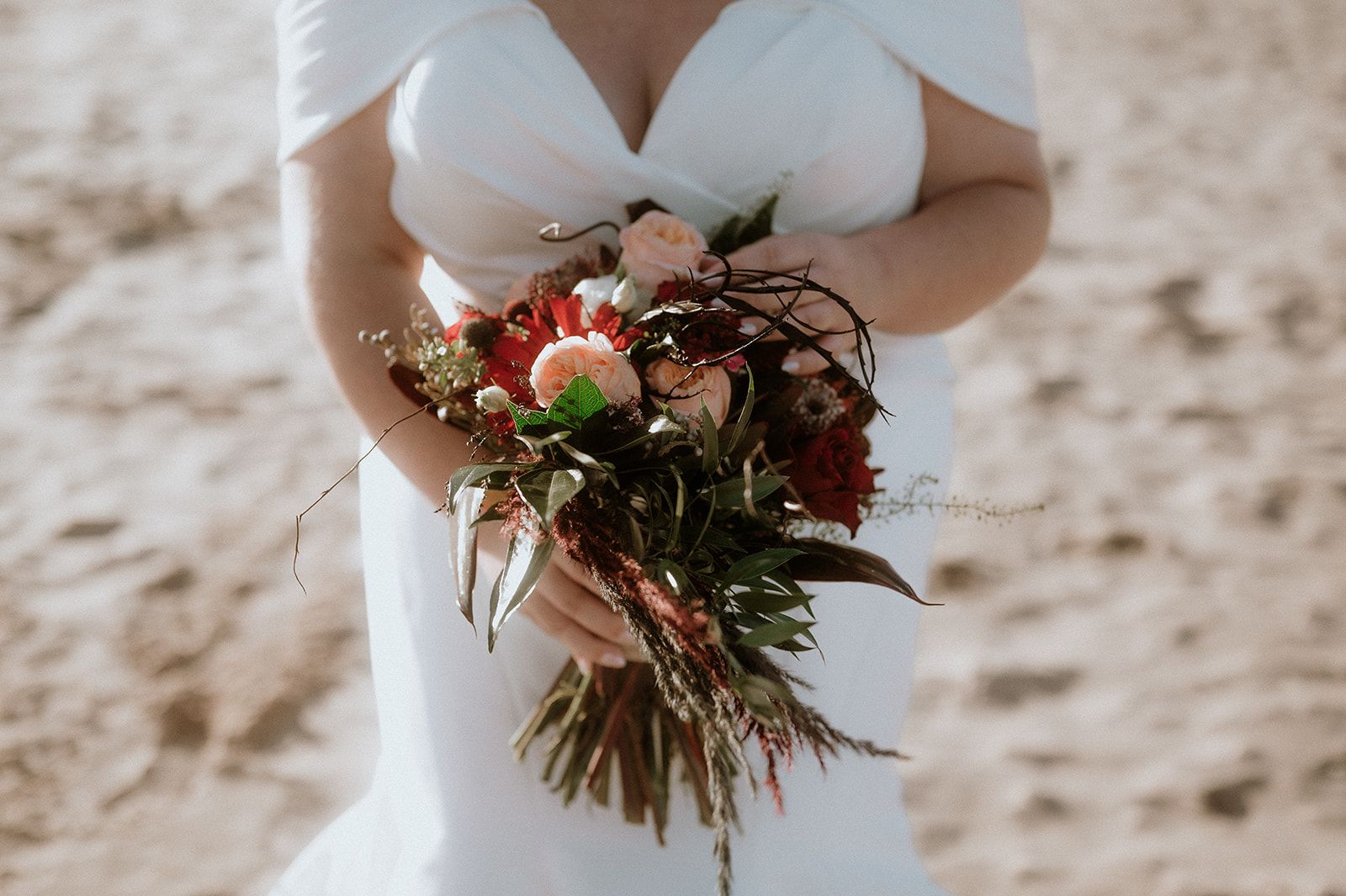 Bride on a beach holding a bouquet of deep red and pink flowers, with some green leaves