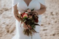 Bride on a beach holding a bouquet of deep red and pink flowers, with some green leaves