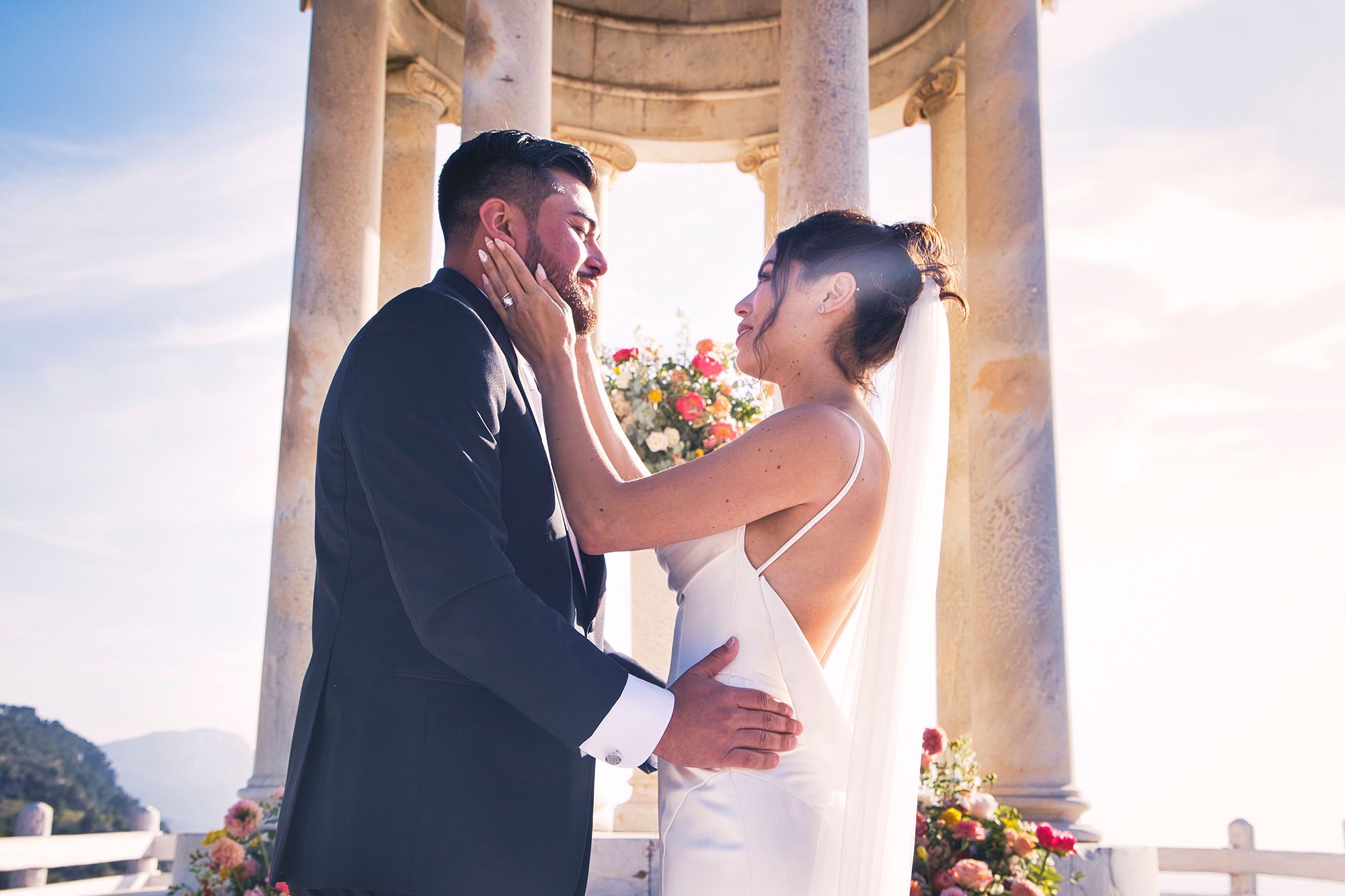 Bride and groom looking at each other with the sun and a stone gazebo behind when they eloped in Spain atop a Mallorcan cliff