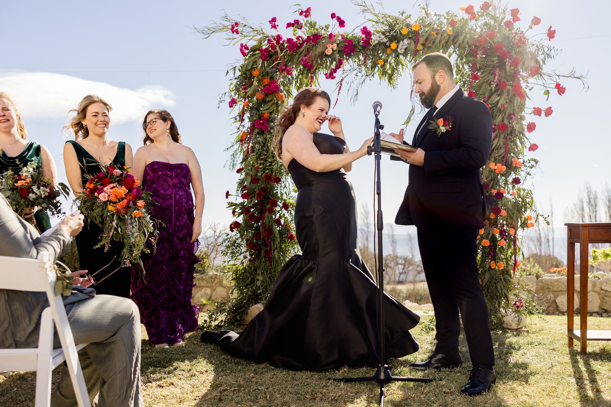 Bride wipes tears while groom recites vows and bridesmaids listen in an outdoor wedding Spain