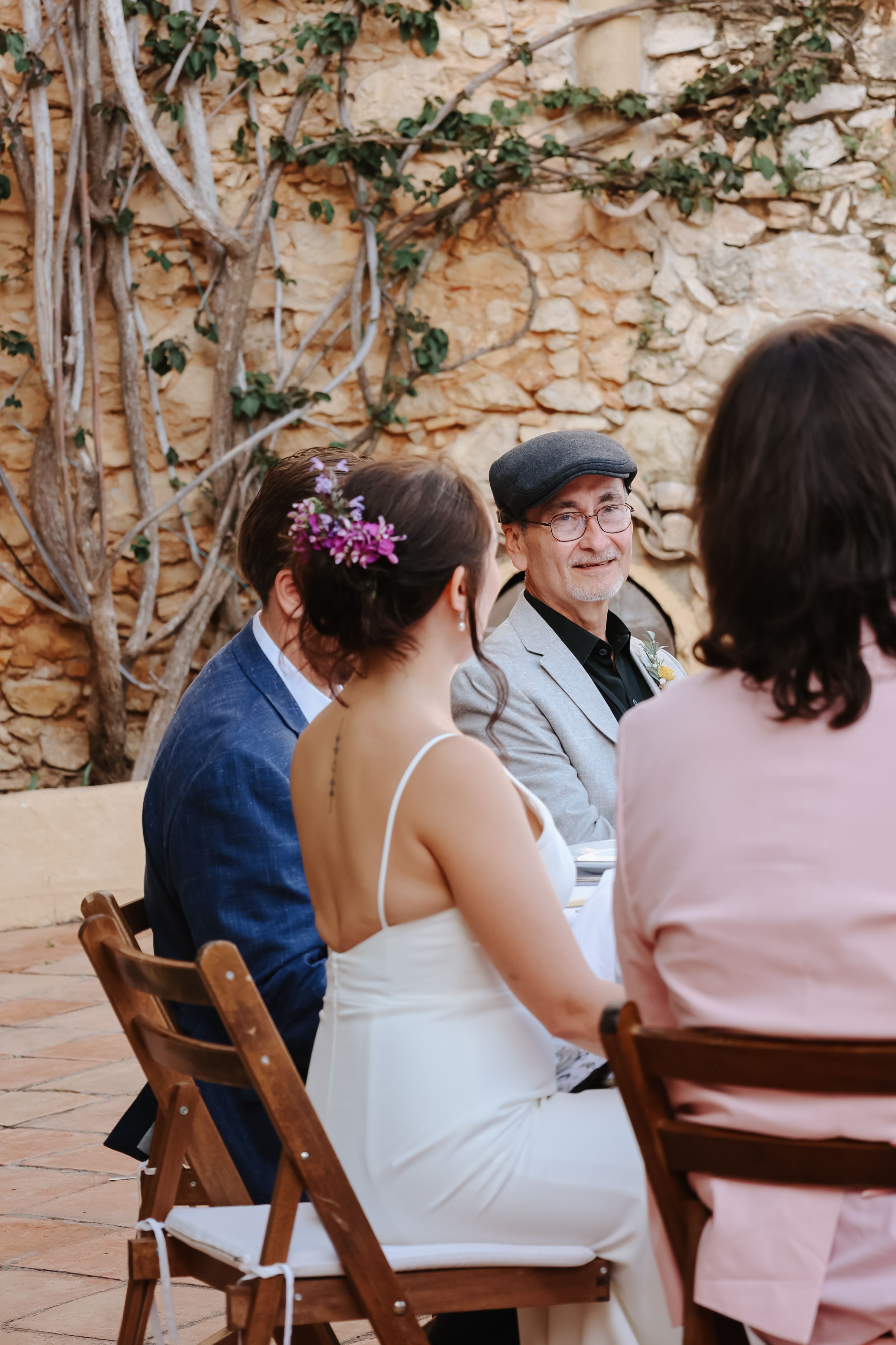 Bride seated with others in a round table with a brick wall in the background and an old man looking at her