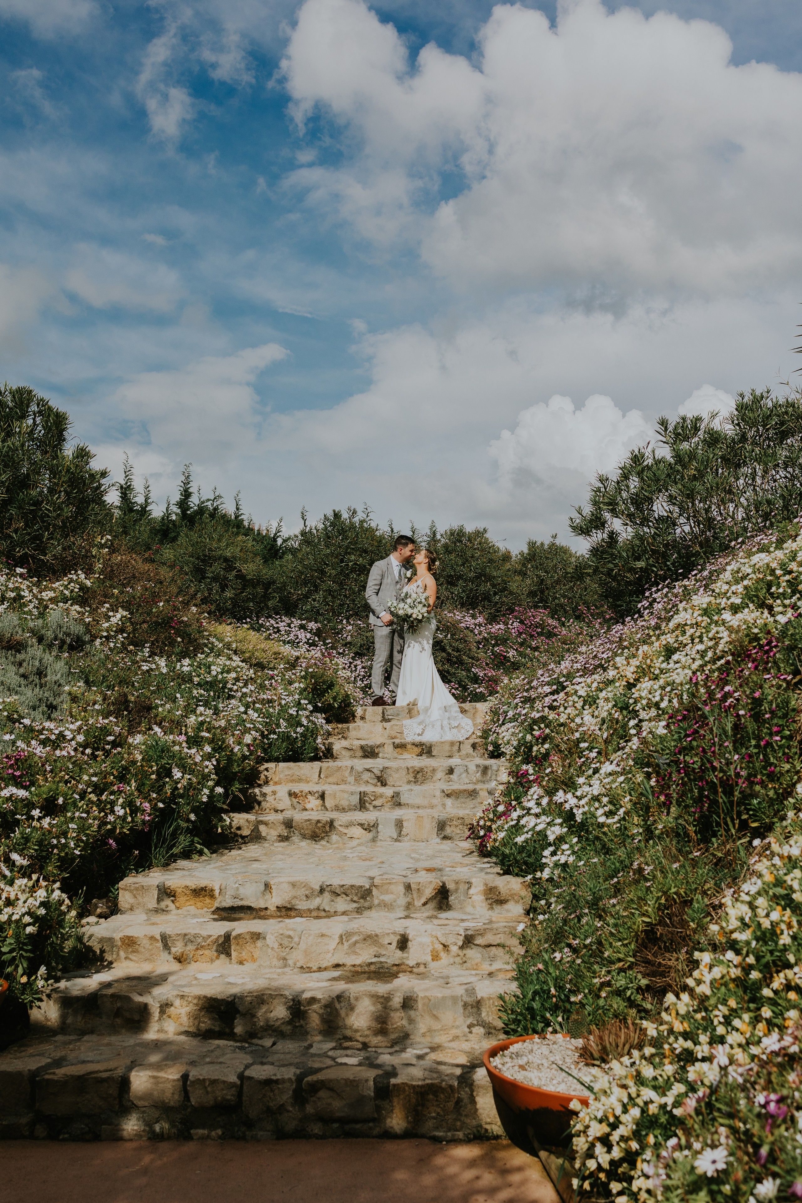 Newlyweds stand atop a staircase surrounded by flower fields during the photoshoot of their small wedding in Portugal