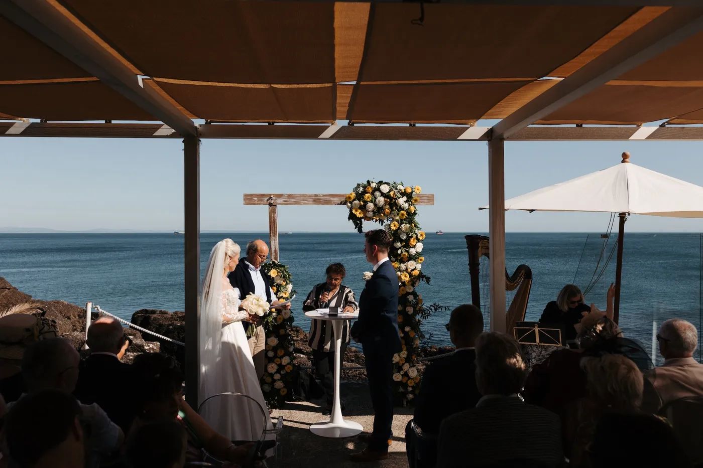 Bride and groom having an outdoor wedding ceremony with a wooden arch decorated with flowers, with ocean in the background