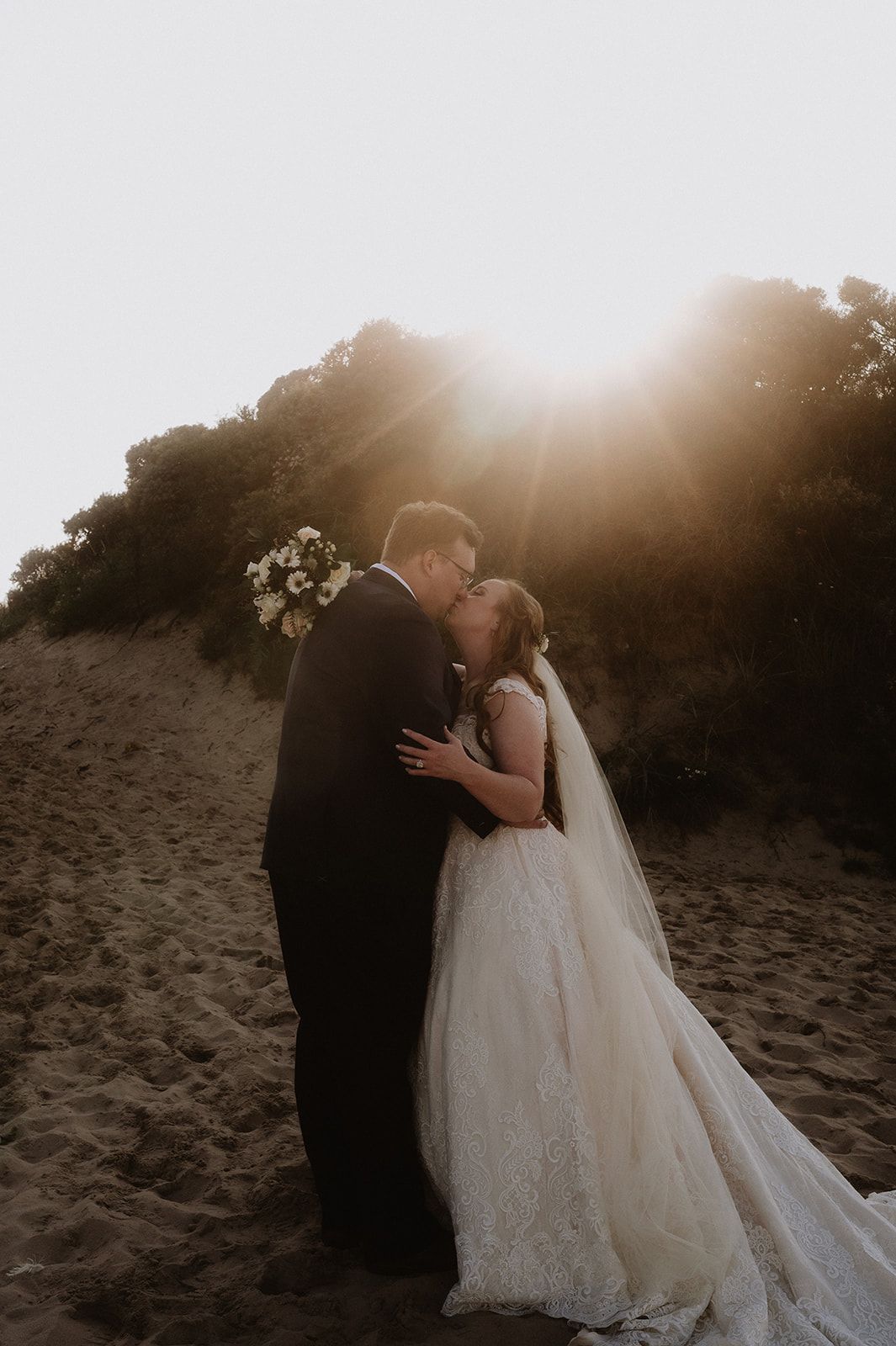 Bride and groom amidst a white-sand beach kiss with sunset behind them during their vow renewal in Ireland