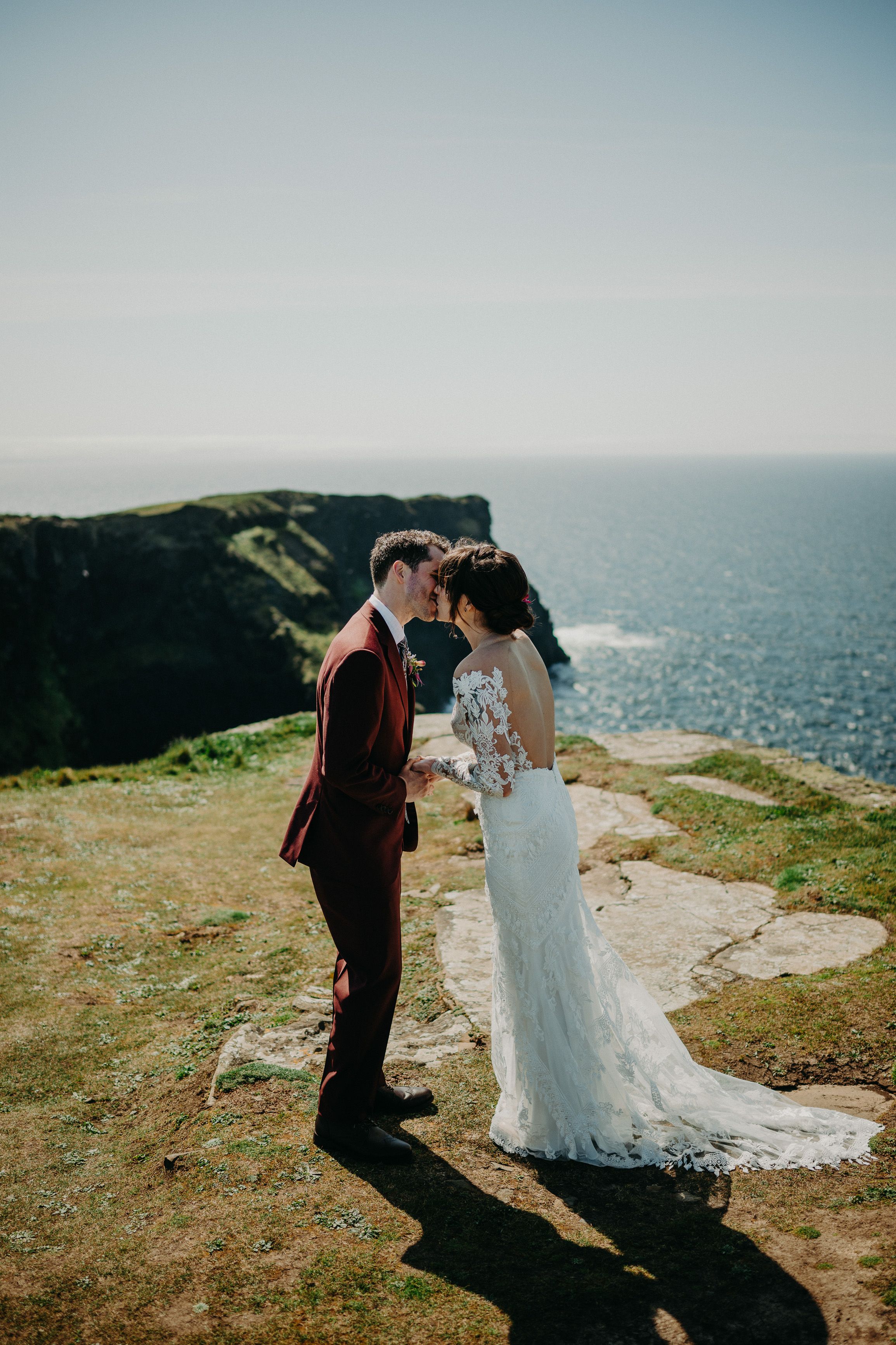 A groom kissing his bride on top of a dramatic cliff with a view of the sea after intimate wedding in Ireland