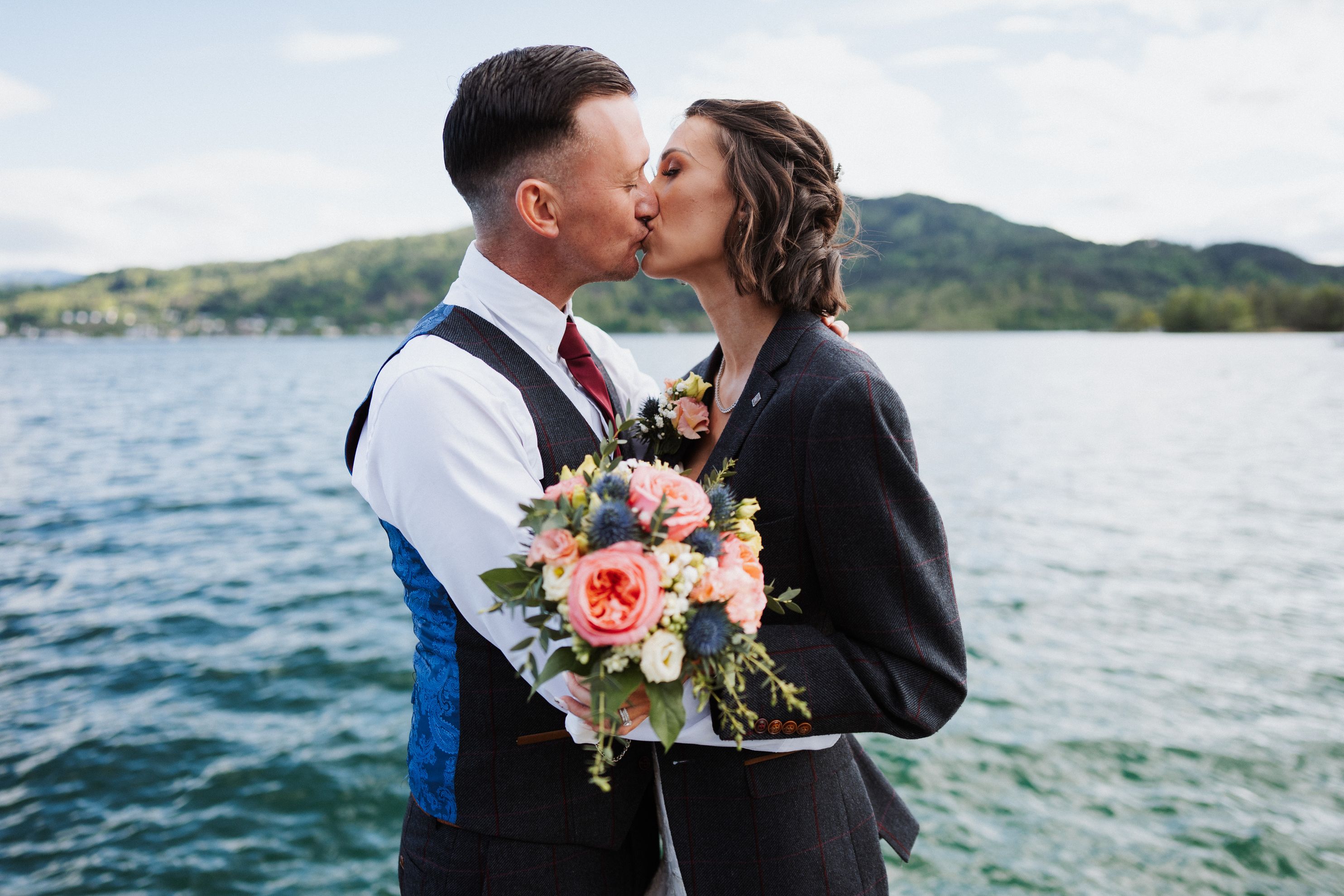 Couple kissing at the water's edge of an Austrian lake, peach bouquet between them