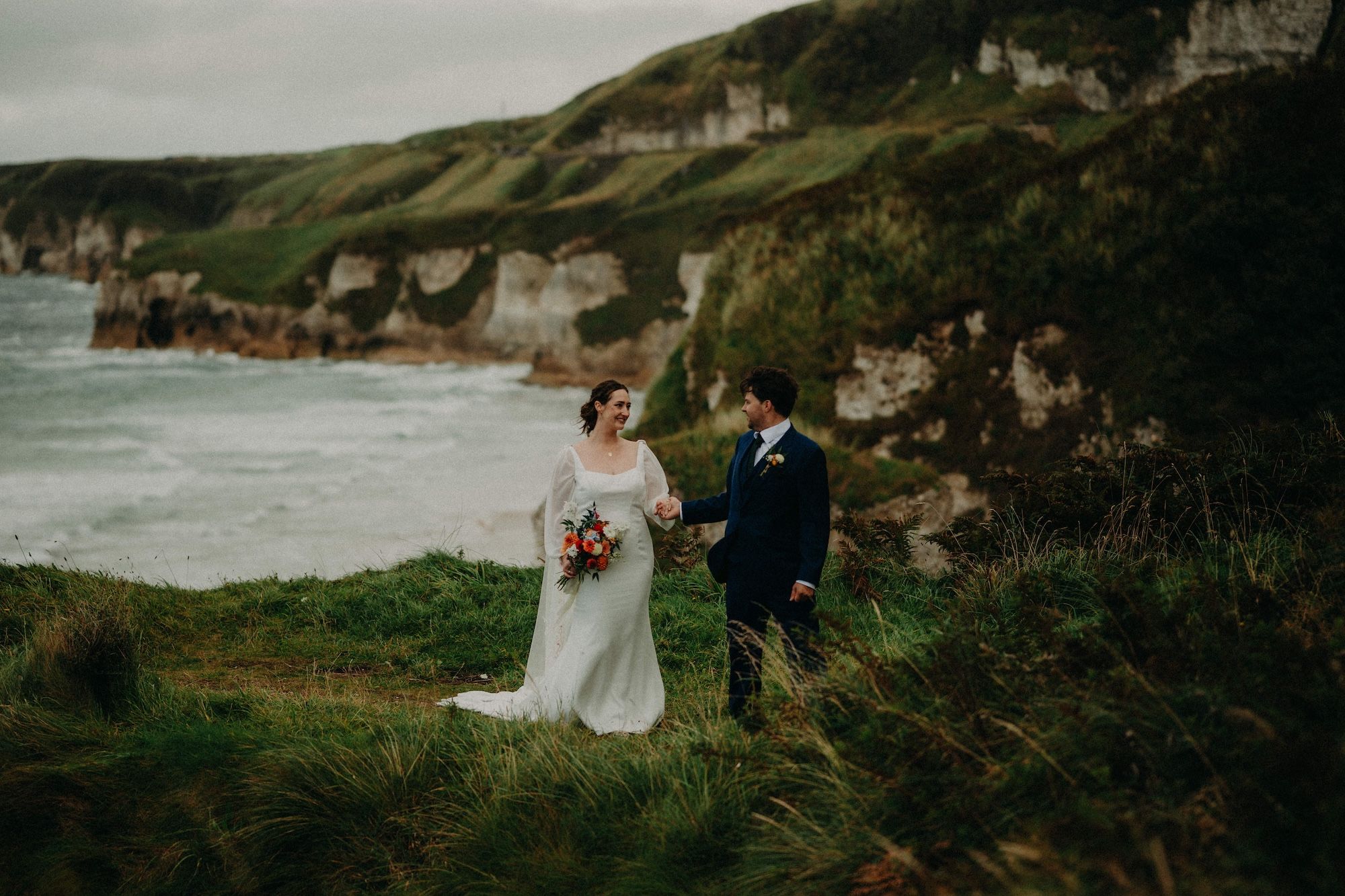 Newlyweds on top of a cliff in Antrim during the photoshoot of their destination wedding in Ireland, with the Atlantic Ocean and cliffs in the background.