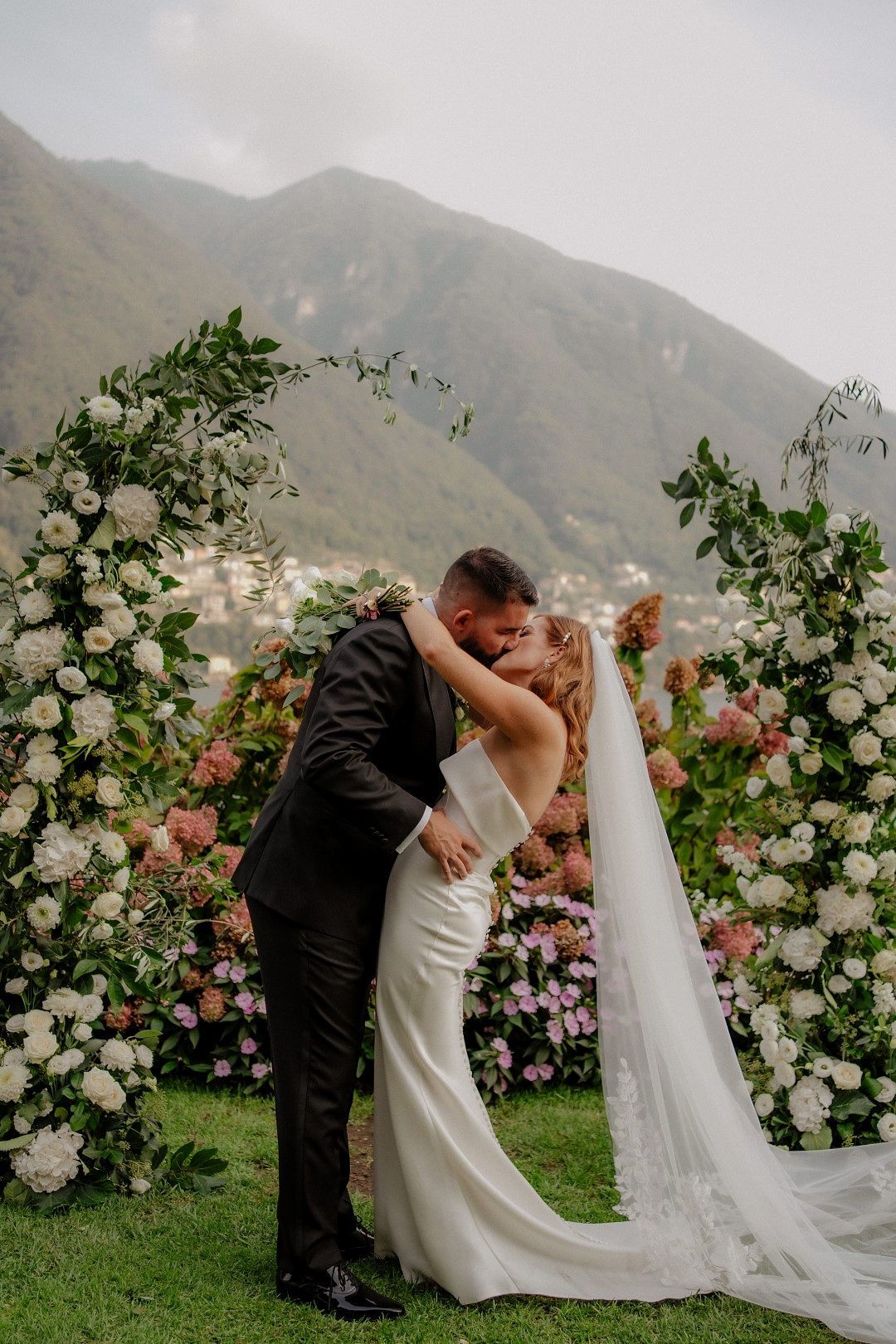 Bride and groom kissing during their intimate wedding ceremony in Italy with a floral arch and Lake Como in the background.