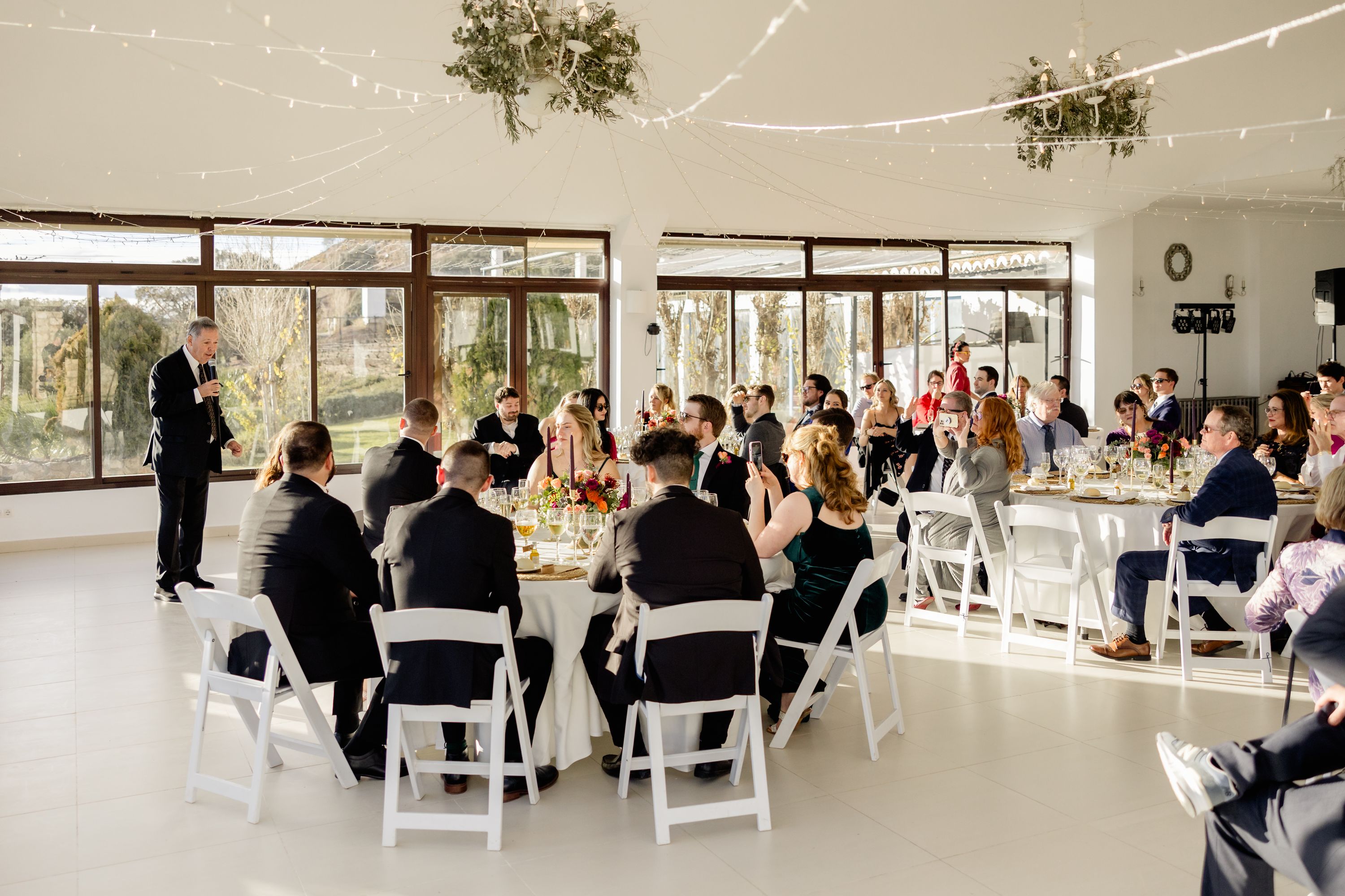 A white reception room filled with tables and chairs and guests listening to someone speaking