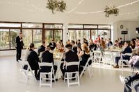 A white reception room filled with tables and chairs and guests listening to someone speaking