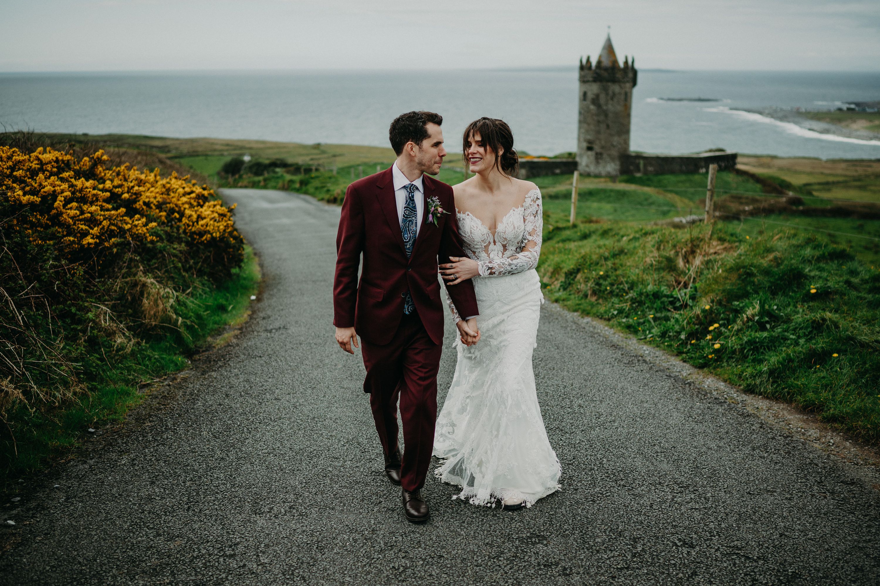 A bride and groom walking down a path with a tower and a view behind them after their small wedding in Ireland